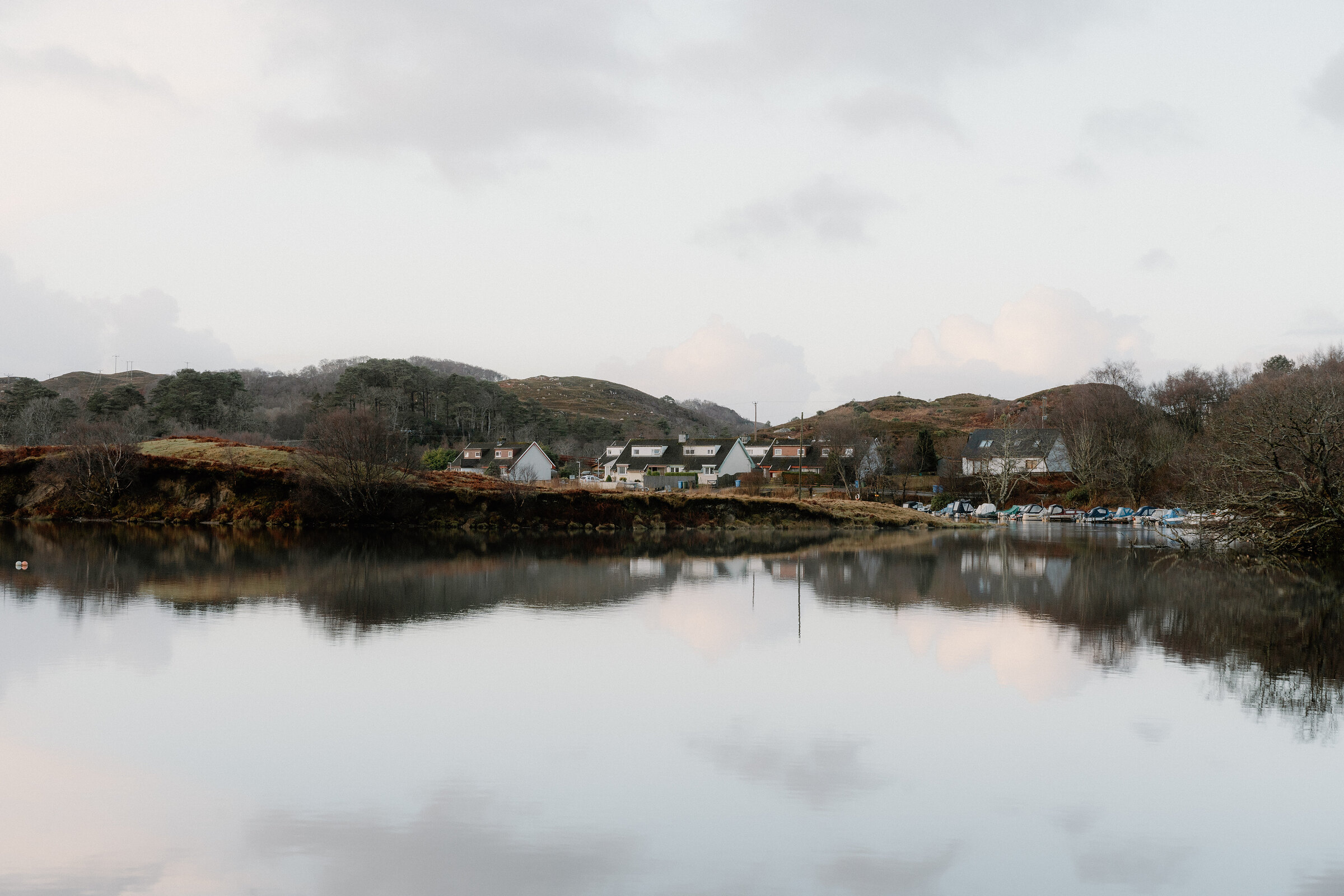 A row of white cottages is mirrored in a still loch, wooded hills rising behind and small boats moored along the shore.