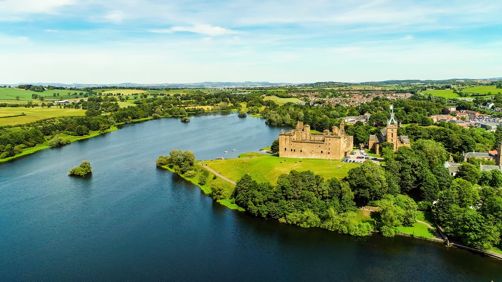Linlithgow Loch from the air on a summer day, the palace and St Michael's Church on its green shore among trees, fields to the horizon.