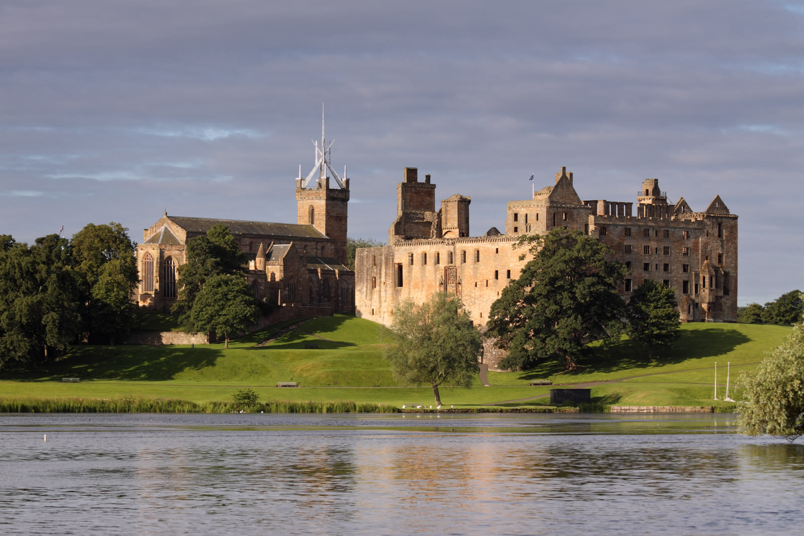 Linlithgow Palace and the crown spire of St Michael's Church across Linlithgow Loch in summer, green grass running down to the water.