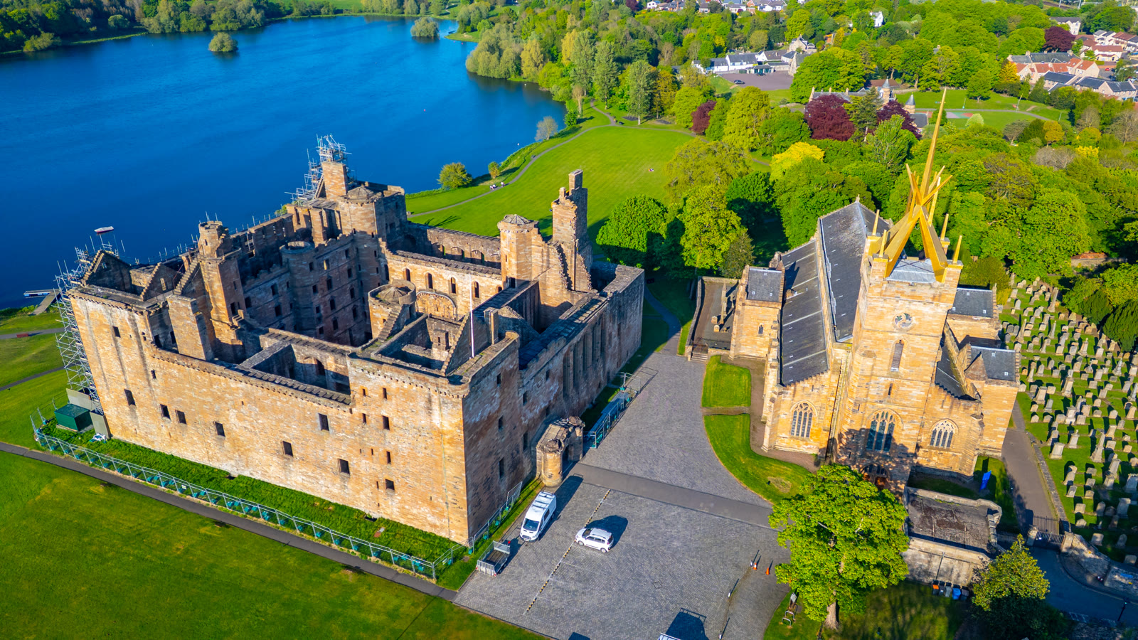Linlithgow Palace from the air, its roofless ranges round the courtyard, St Michael's Church and its crown spire beside it and the loch beyond.