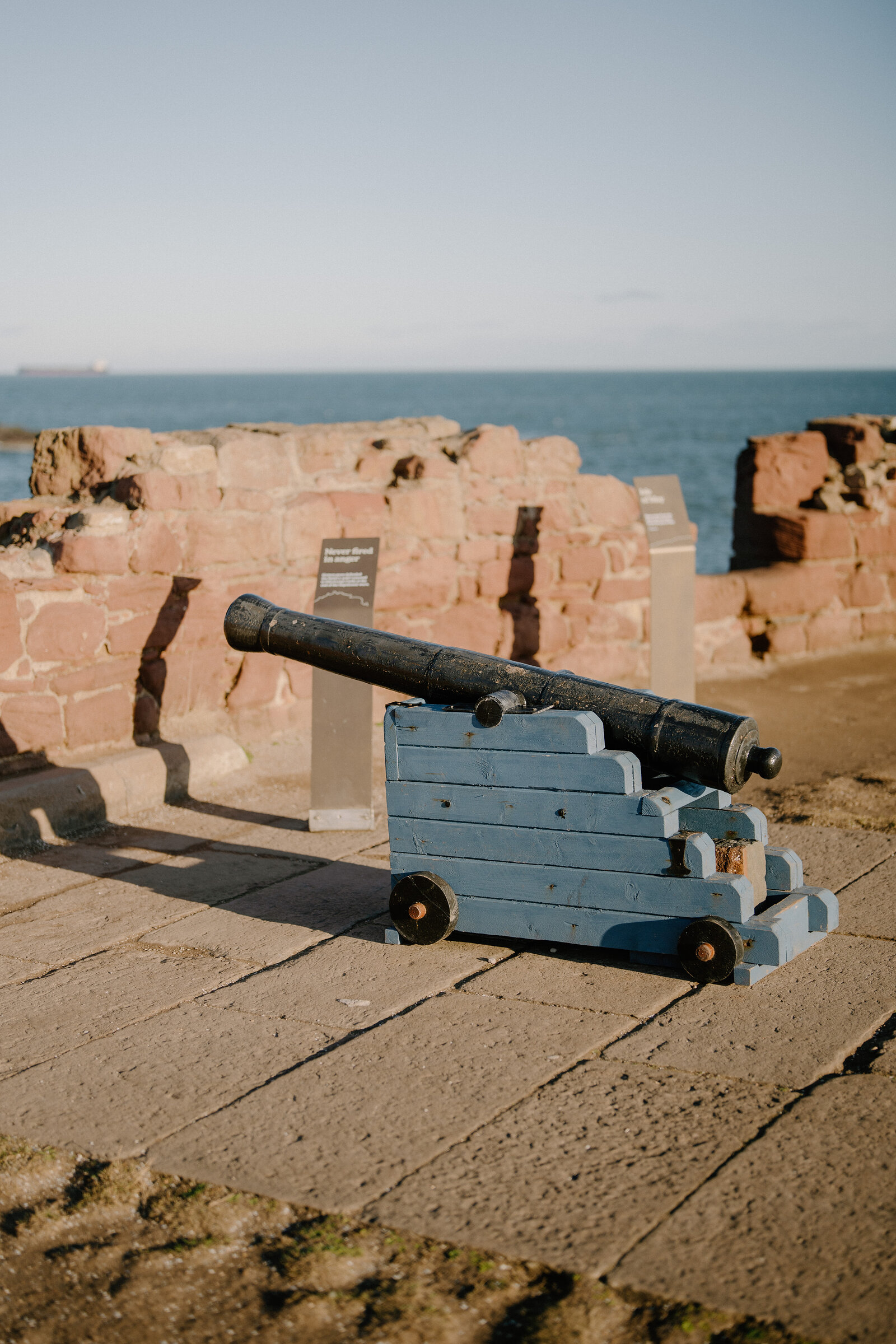 A cannon on a wooden carriage points out to sea beside the ruined red sandstone walls of a castle, information panels standing nearby.