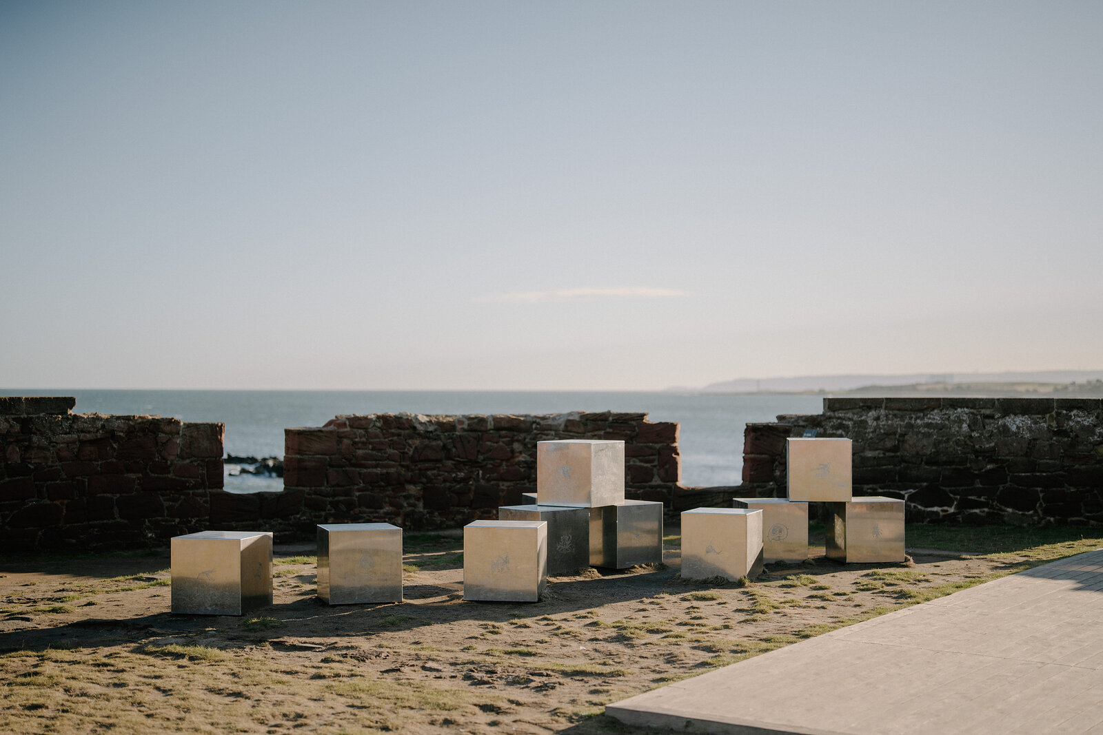 A cluster of reflective metal cubes stands arranged on gravel within the ruined walls of a castle, the sea visible beyond the battlements.