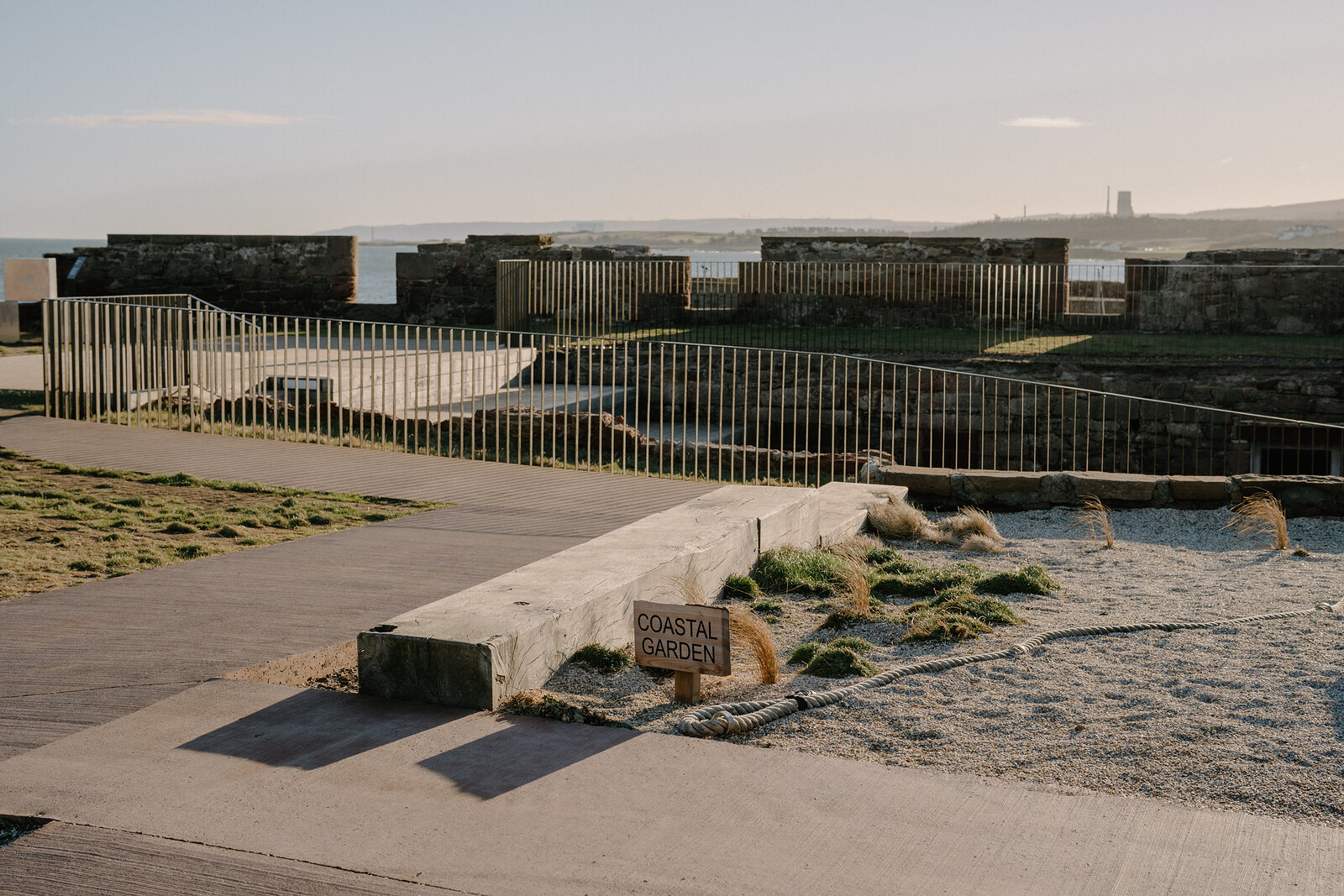A sign reading "Coastal Garden" stands among railings and raised beds within the ruined walls of a castle, a town visible on the headland beyond.