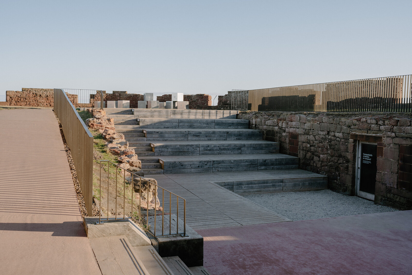 Wide stone steps descend through the ruined walls of a castle terrace, a modern glass-fronted building standing at the base.