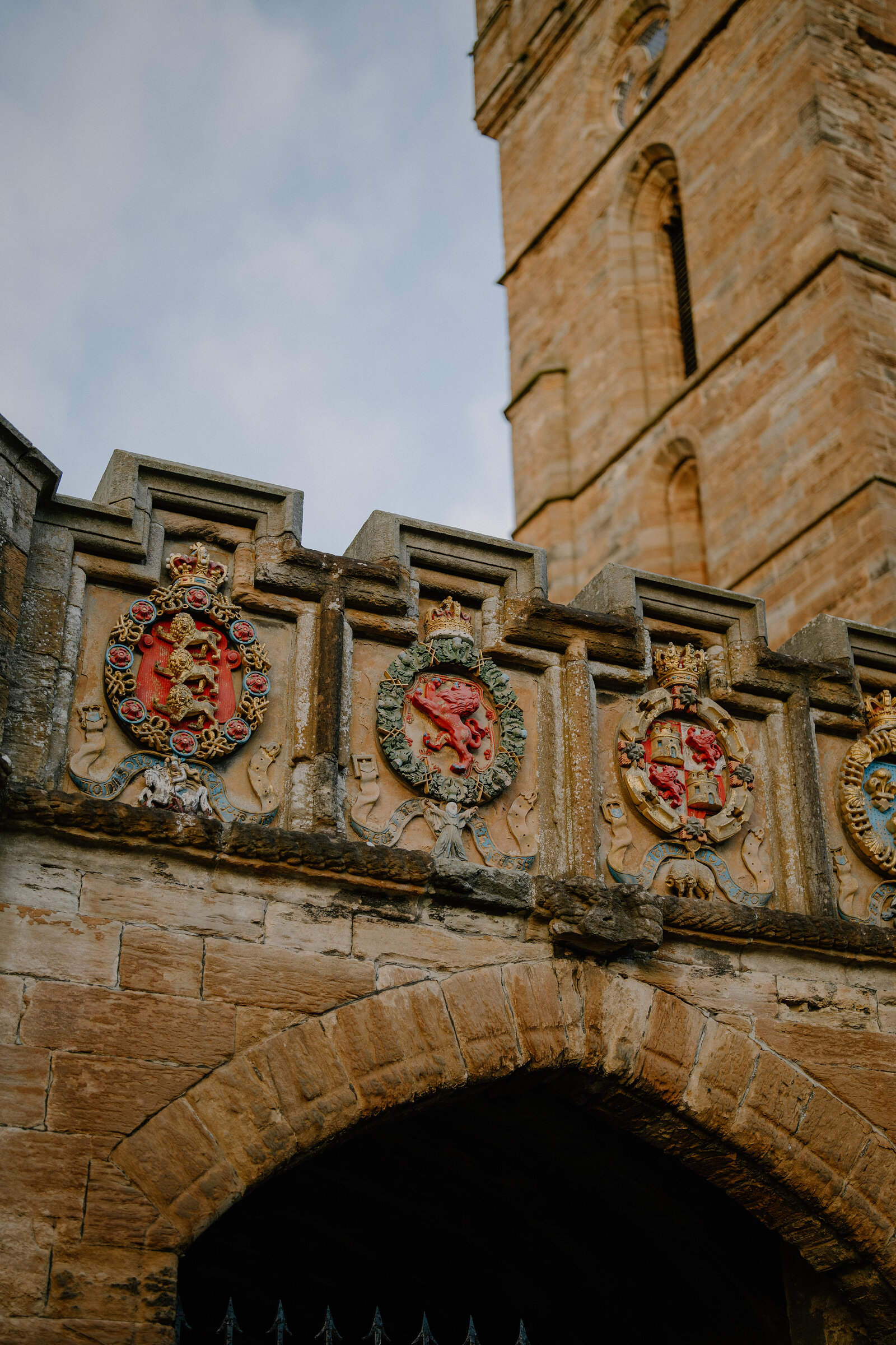Painted heraldic shields carved into a stone parapet line the top of an arched gateway beneath a sandstone church tower.
