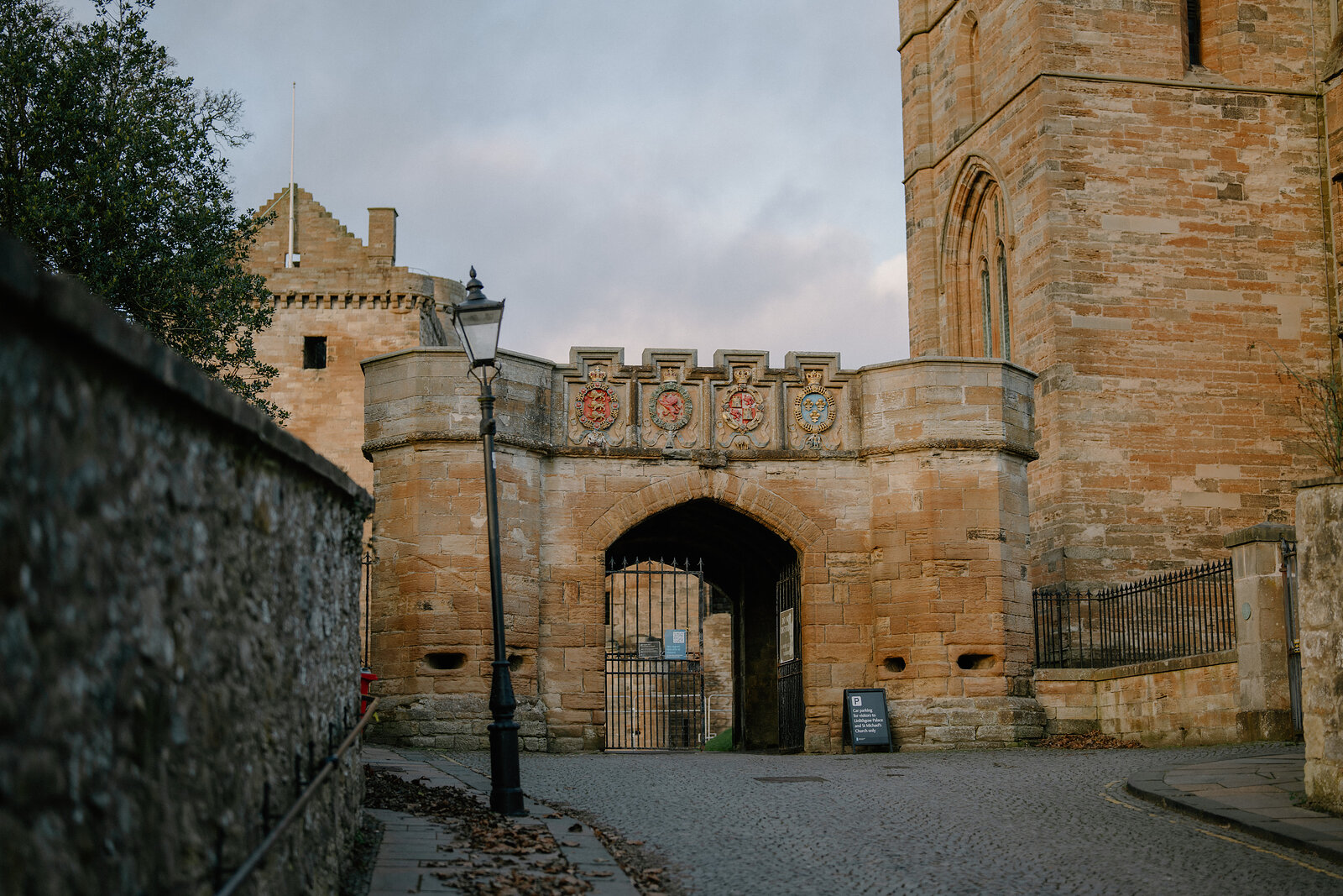 Painted heraldic shields line the top of a stone rampart wall with an arched gateway, a lamp post and church tower rising behind.