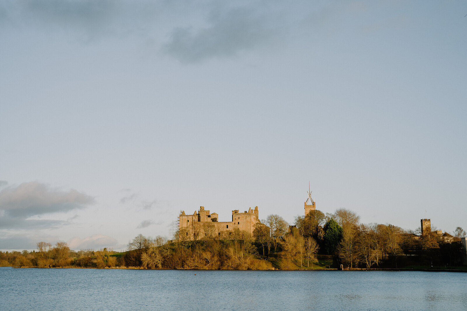 A castle with towers and a domed tower stands on a wooded peninsula reflected in a calm loch at sunset.