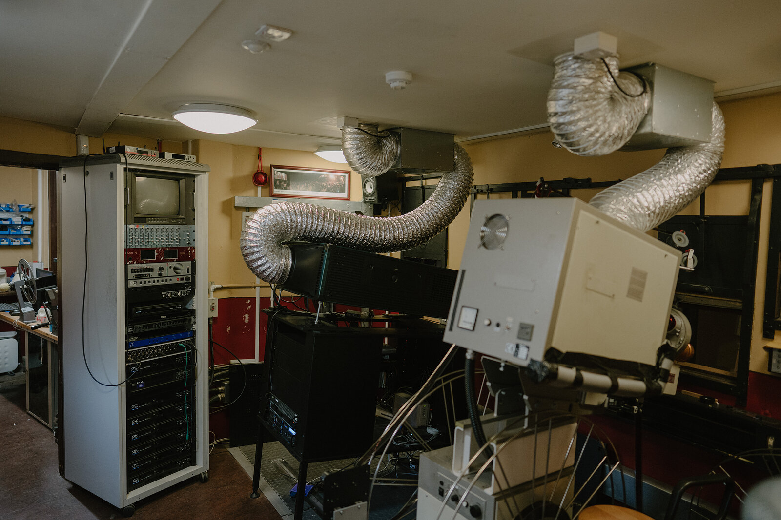 An old film projector with ventilation ducting stands in a cinema projection room beside a rack of vintage equipment.