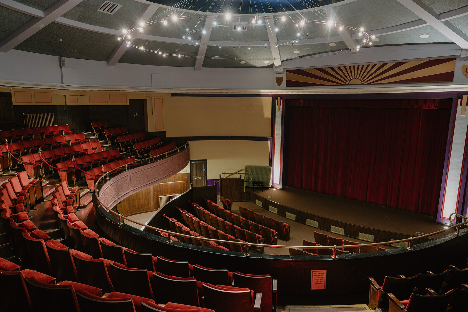 A theatre auditorium with a curved balcony and rows of red velvet seats faces a stage framed by a sunburst-patterned proscenium and dark curtain.