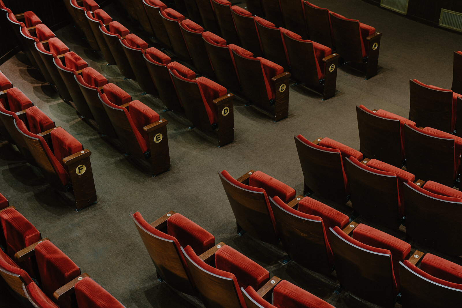 Rows of empty red velvet theatre seats with numbered end panels are seen from above, dividing into two curved sections either side of an aisle.