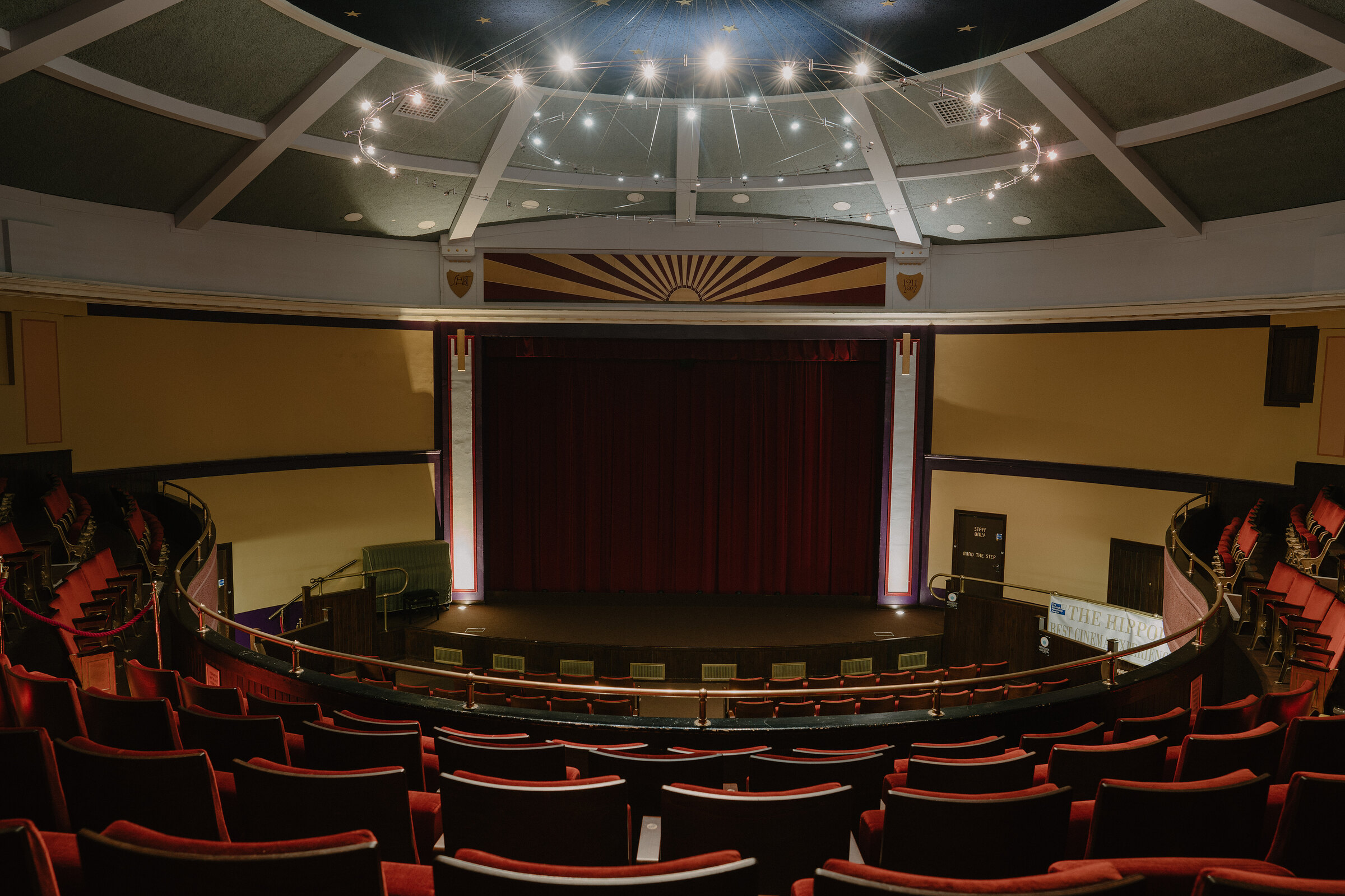 A domed theatre ceiling with rings of lights frames a sunburst-painted proscenium above a red curtain and empty stage, with red seats curving round the auditorium below.