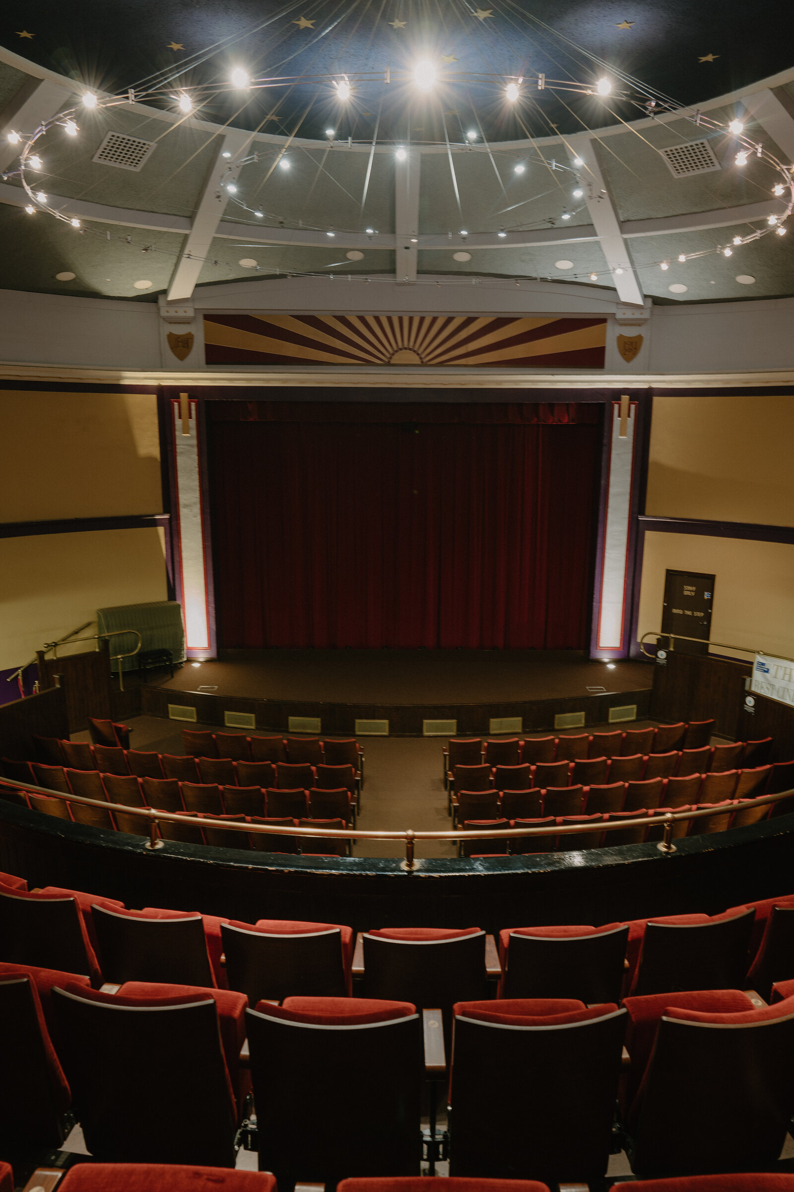 A domed theatre ceiling strung with lights frames a sunburst-painted proscenium above a closed red stage curtain, with rows of red seats facing it.