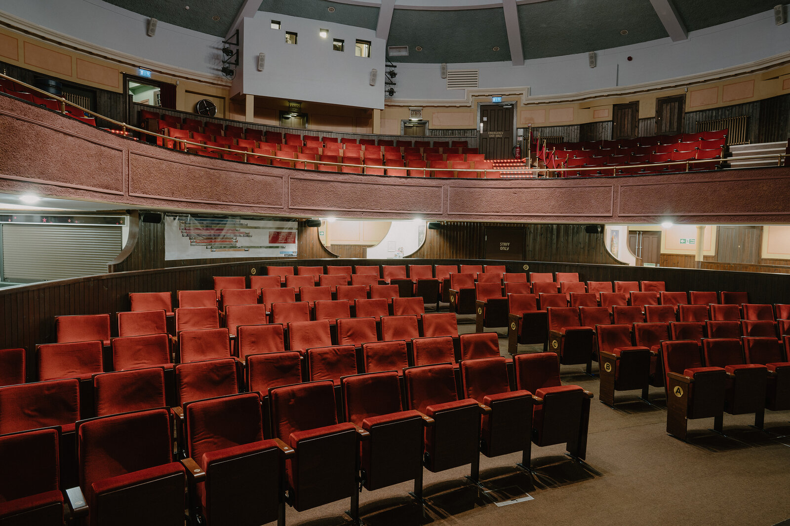 A curved theatre balcony with rows of red seats overlooks the stalls below, seen from the rear of the upper level under a domed ceiling.