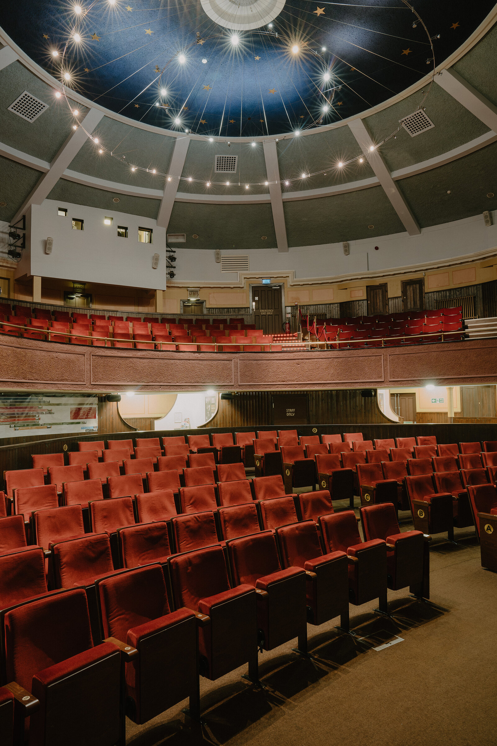 A domed theatre ceiling painted with stars and strung with lights rises above a curved balcony of red seats and a lower tier of seating below.