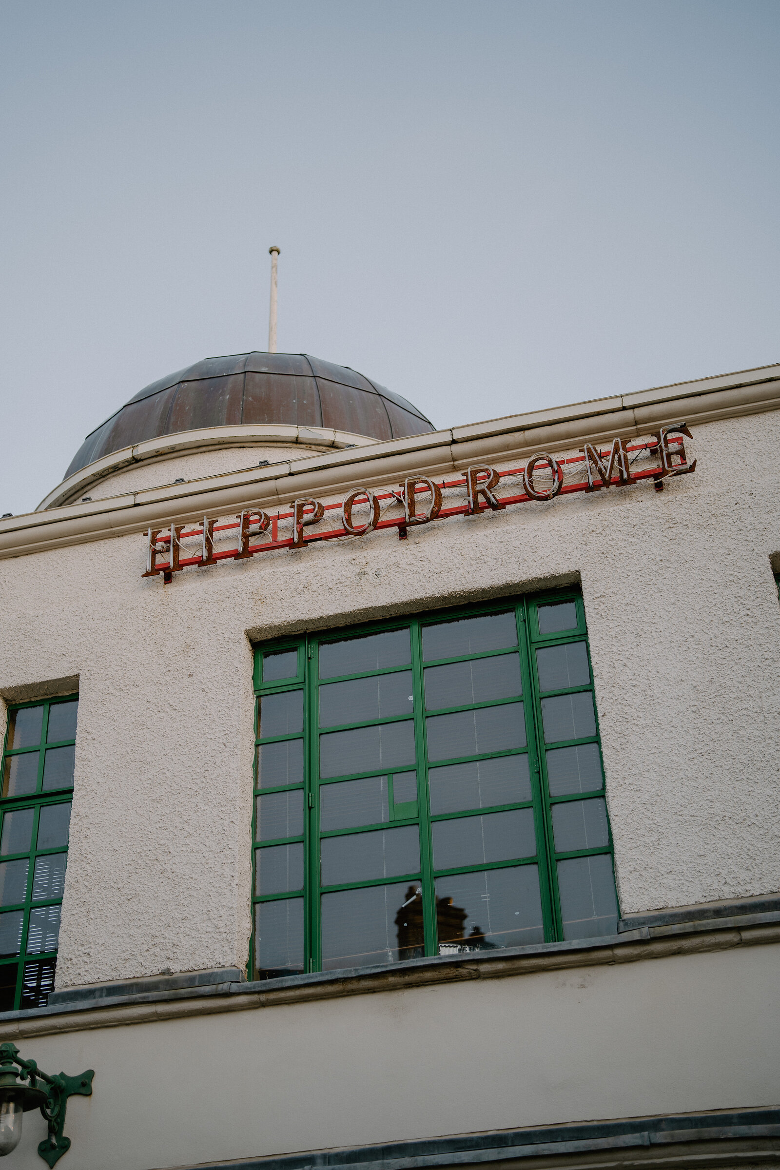 Red neon letters spelling "Hippodrome" run below a domed roof on a cream-rendered building against an overcast sky.
