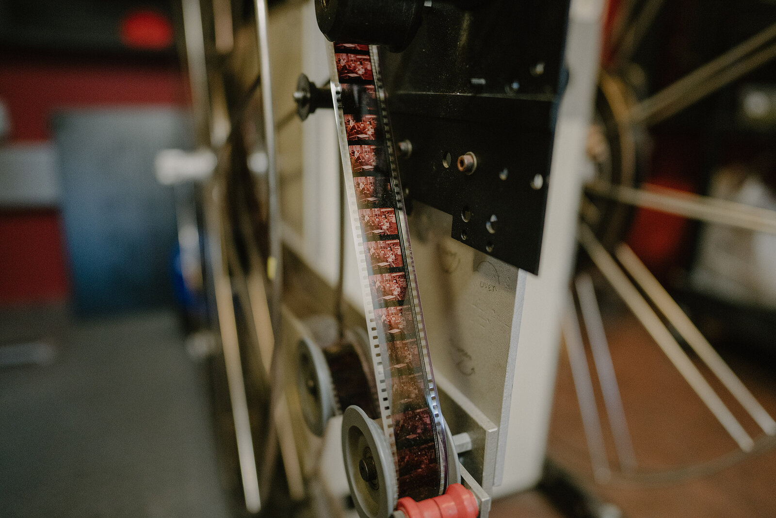 A strip of exposed film runs through a projector's sprockets and reels in close-up, red light reflecting off the machine.
