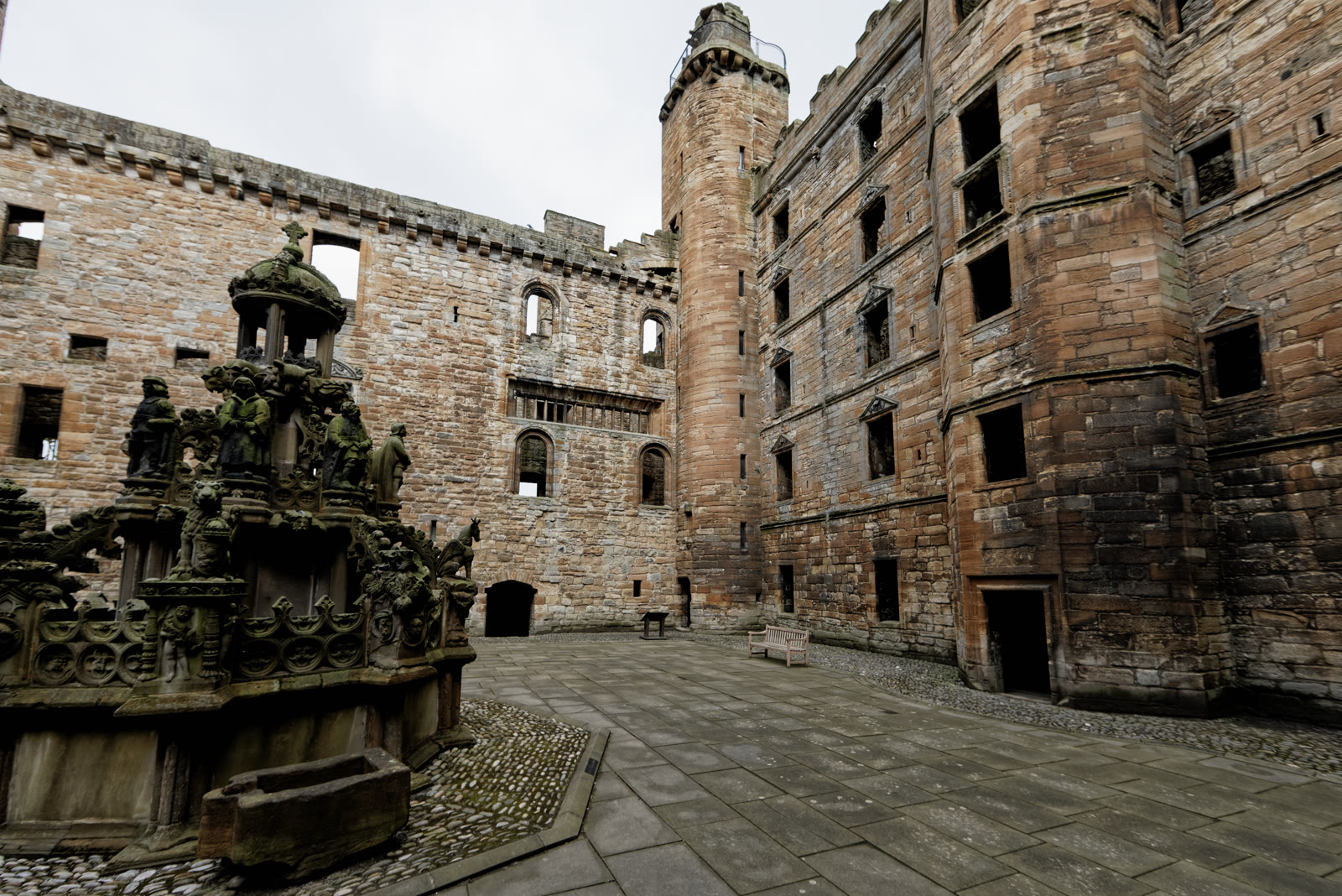 The carved stone fountain in the courtyard of Linlithgow Palace, roofless sandstone ranges and a stair tower standing round it.