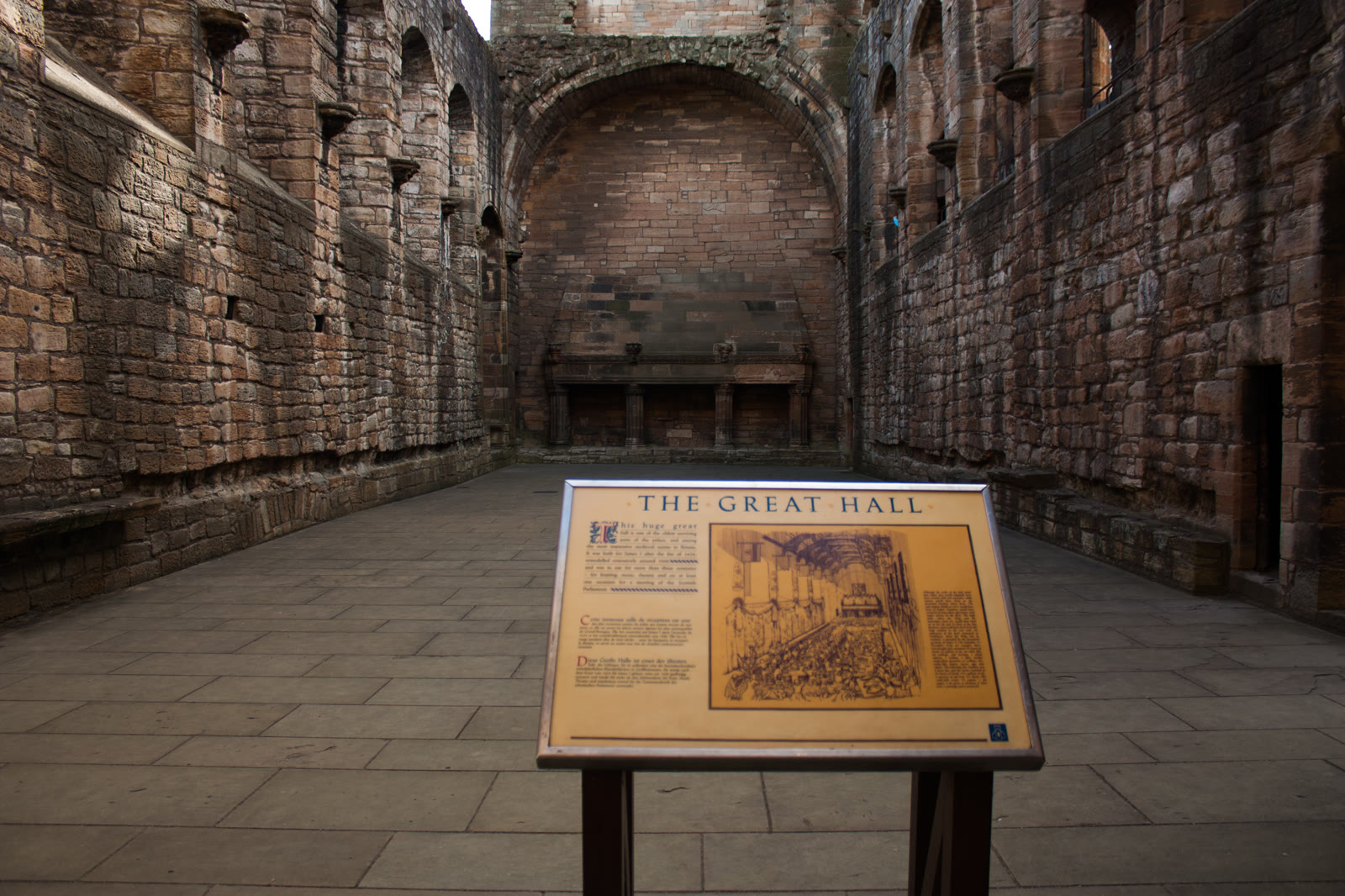 The roofless great hall of Linlithgow Palace, long sandstone walls running to an arched recess at the far end, a board in front reading The Great Hall.