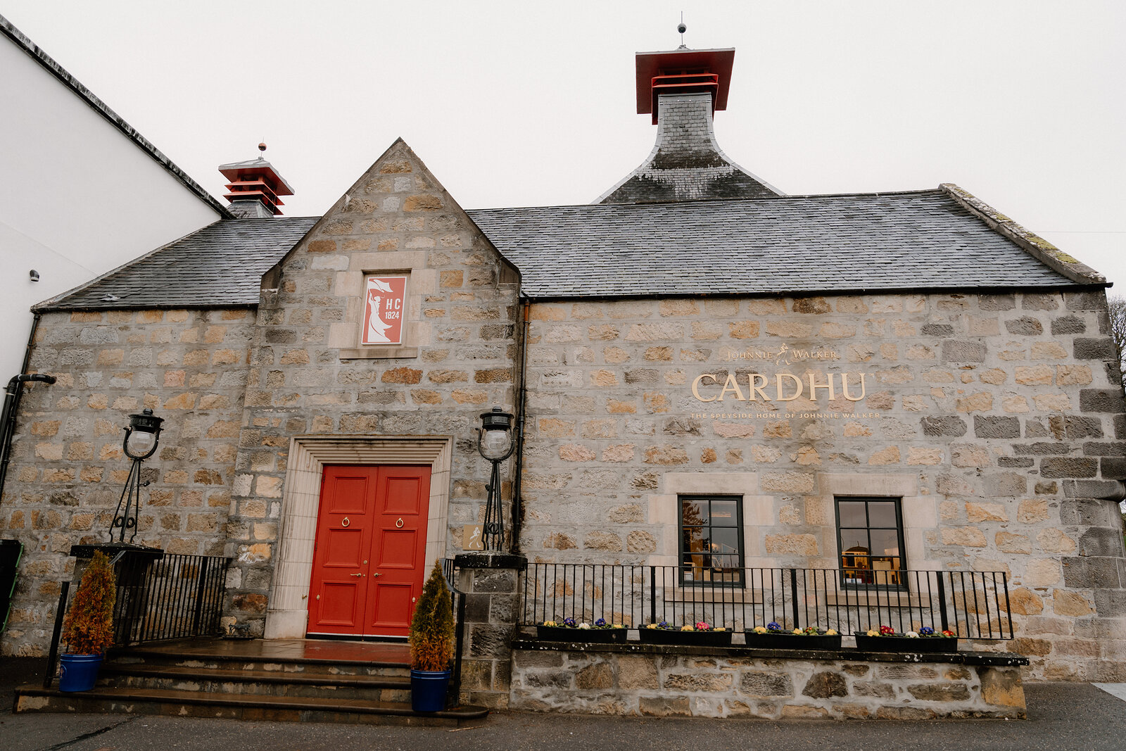 Cardhu's stone distillery building is lettered above a red door flanked by potted plants.