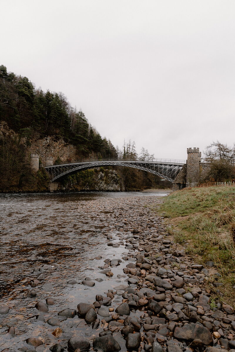 Craigellachie Bridge crosses the River Spey below wooded cliffs, seen from its stony bank.