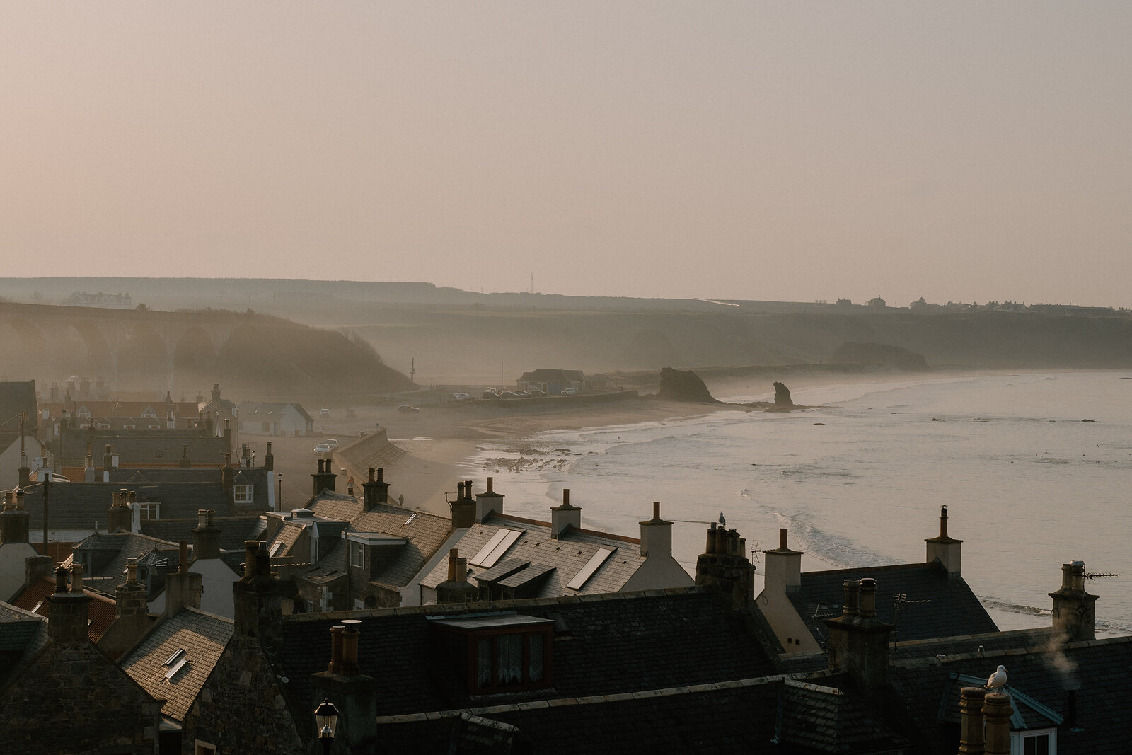 Rooftops and chimneys of a Moray coastal town overlook a beach curving toward distant sea stacks.