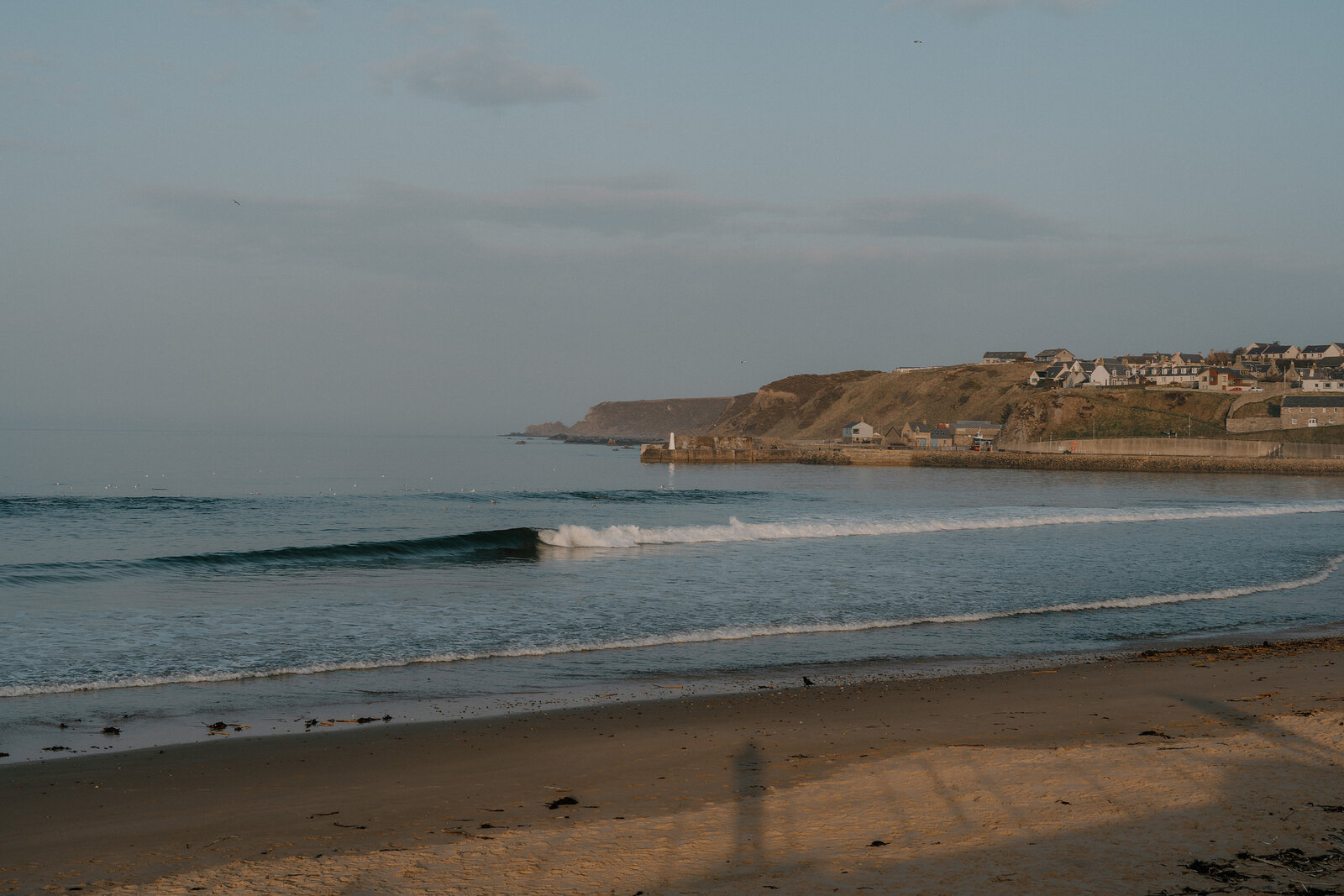 Small waves break on a sandy Moray beach beneath cliffs and clustered houses.
