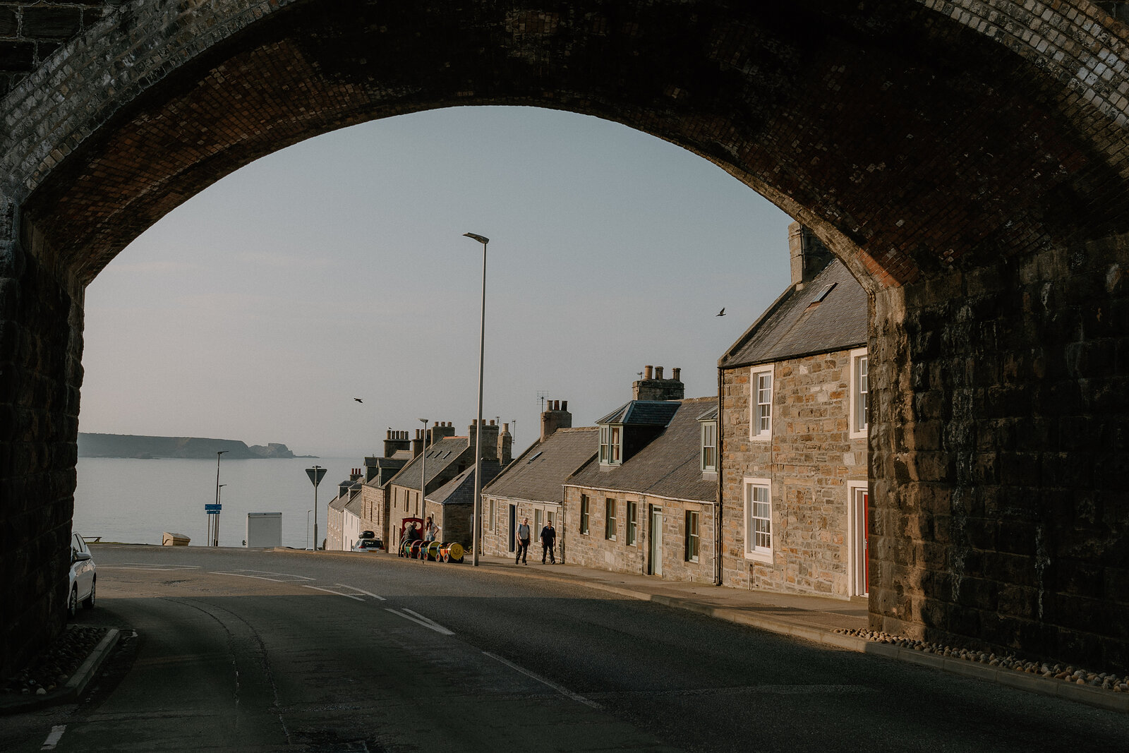 A stone railway viaduct arch frames a row of houses and the sea beyond on a Moray seafront street.