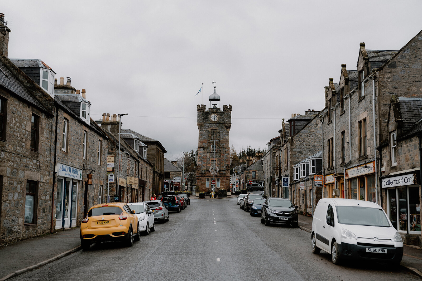 Parked cars line a Dufftown street leading toward the castellated clock tower beneath a grey sky.