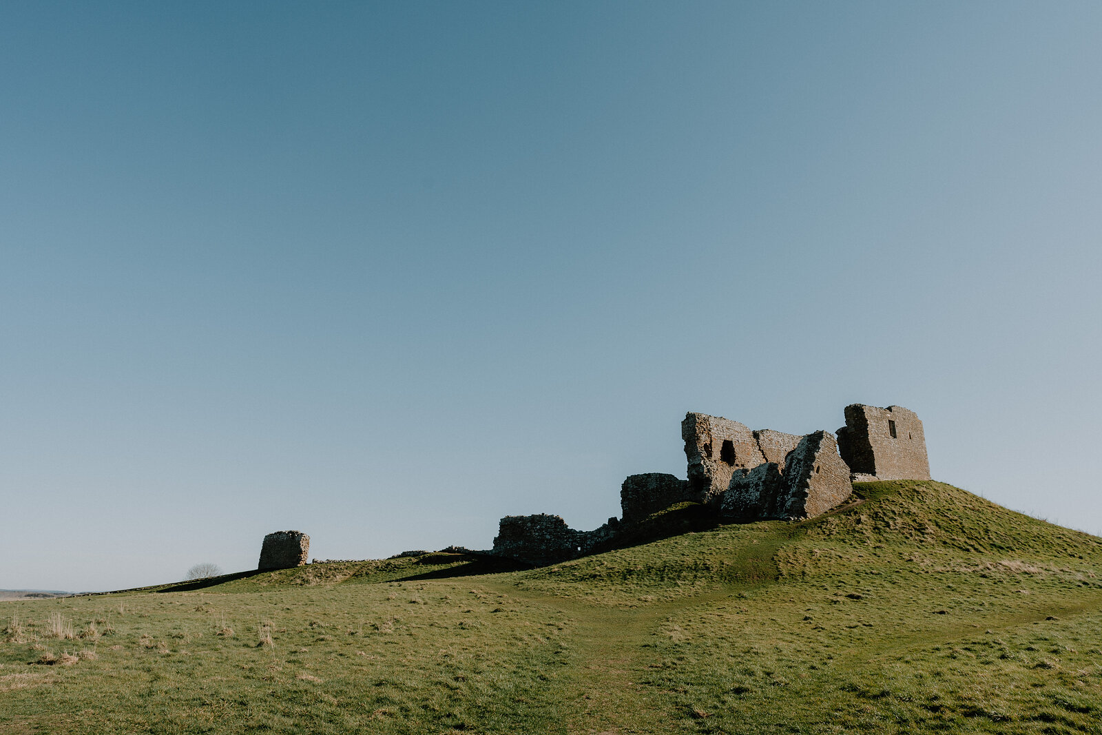 Duffus Castle's ruined keep and a separate stone tower rise from a grassy hillside beneath a clear sky.