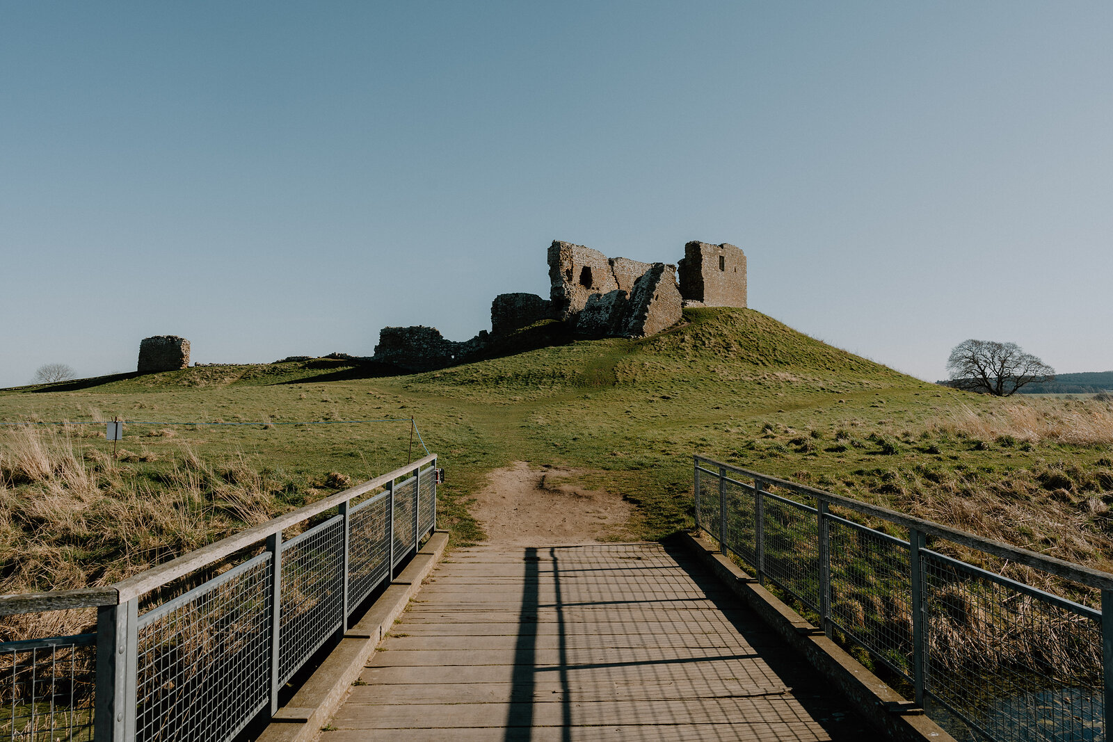 A wooden footbridge leads toward Duffus Castle's ruined stone keep on its grassy motte.