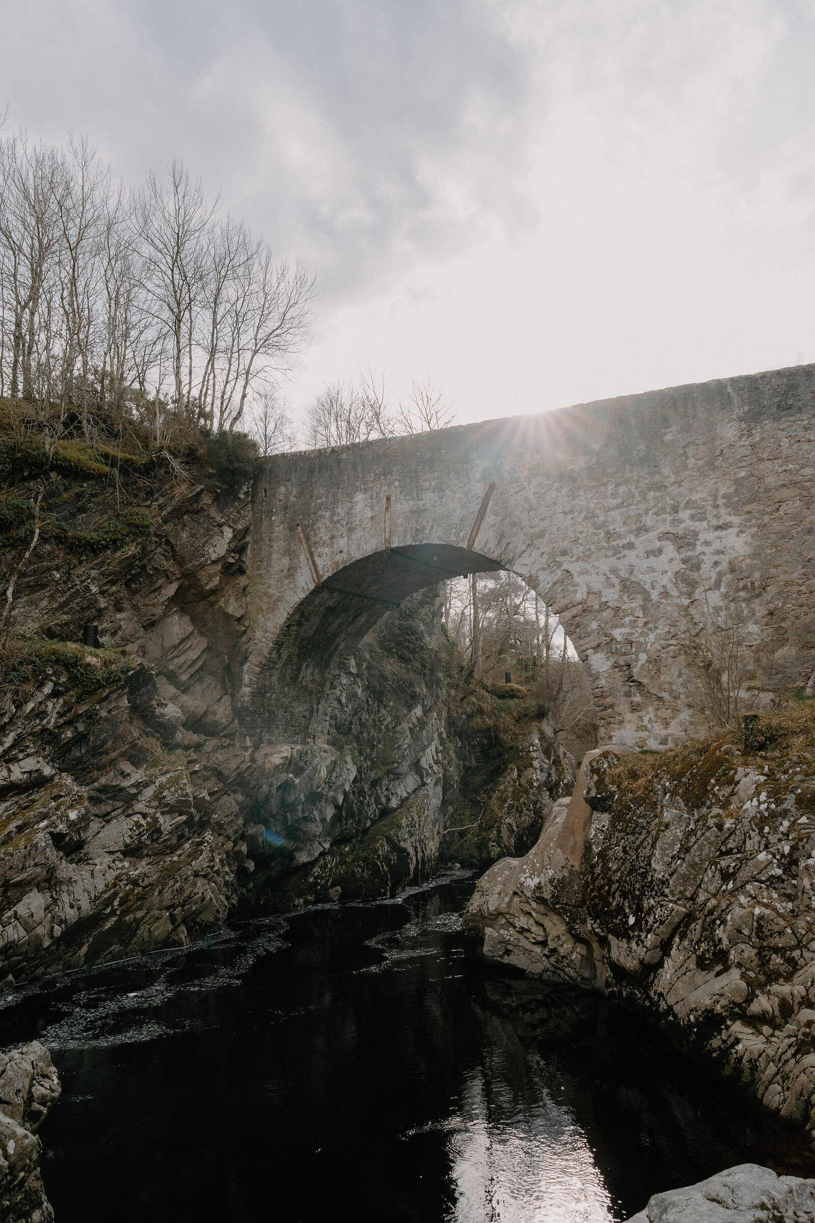 A stone arched bridge spans a rocky river gorge, lit by low sun through bare trees.