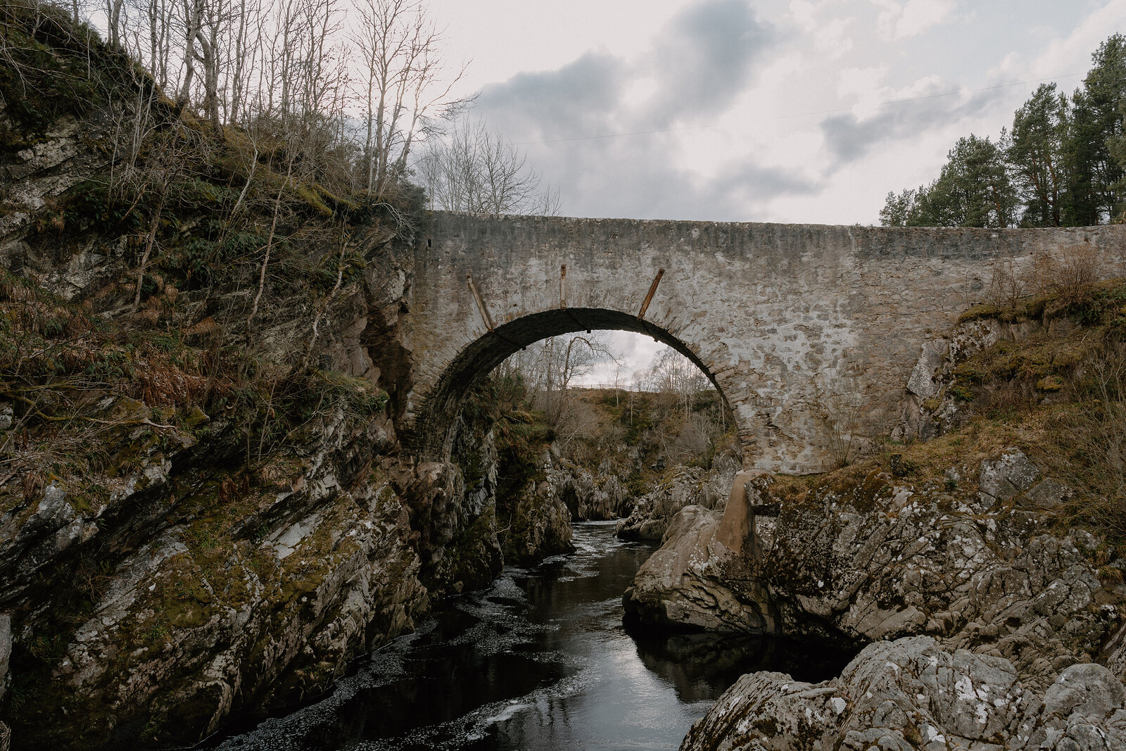 A stone arched bridge spans a river gorge flanked by moss-covered rocks in Moray.