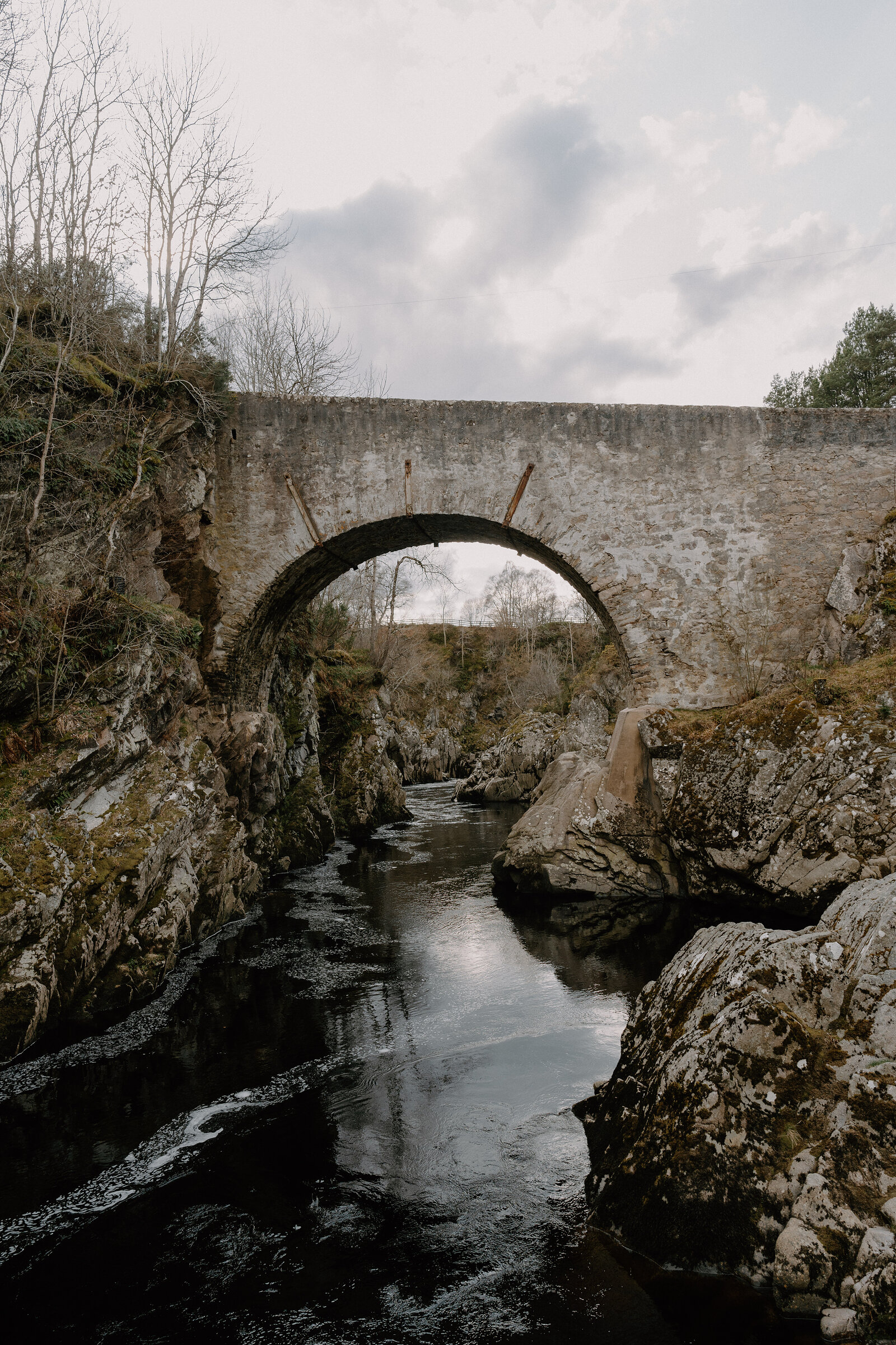 A stone arched bridge spans a dark river gorge between moss-covered rock walls in Moray.