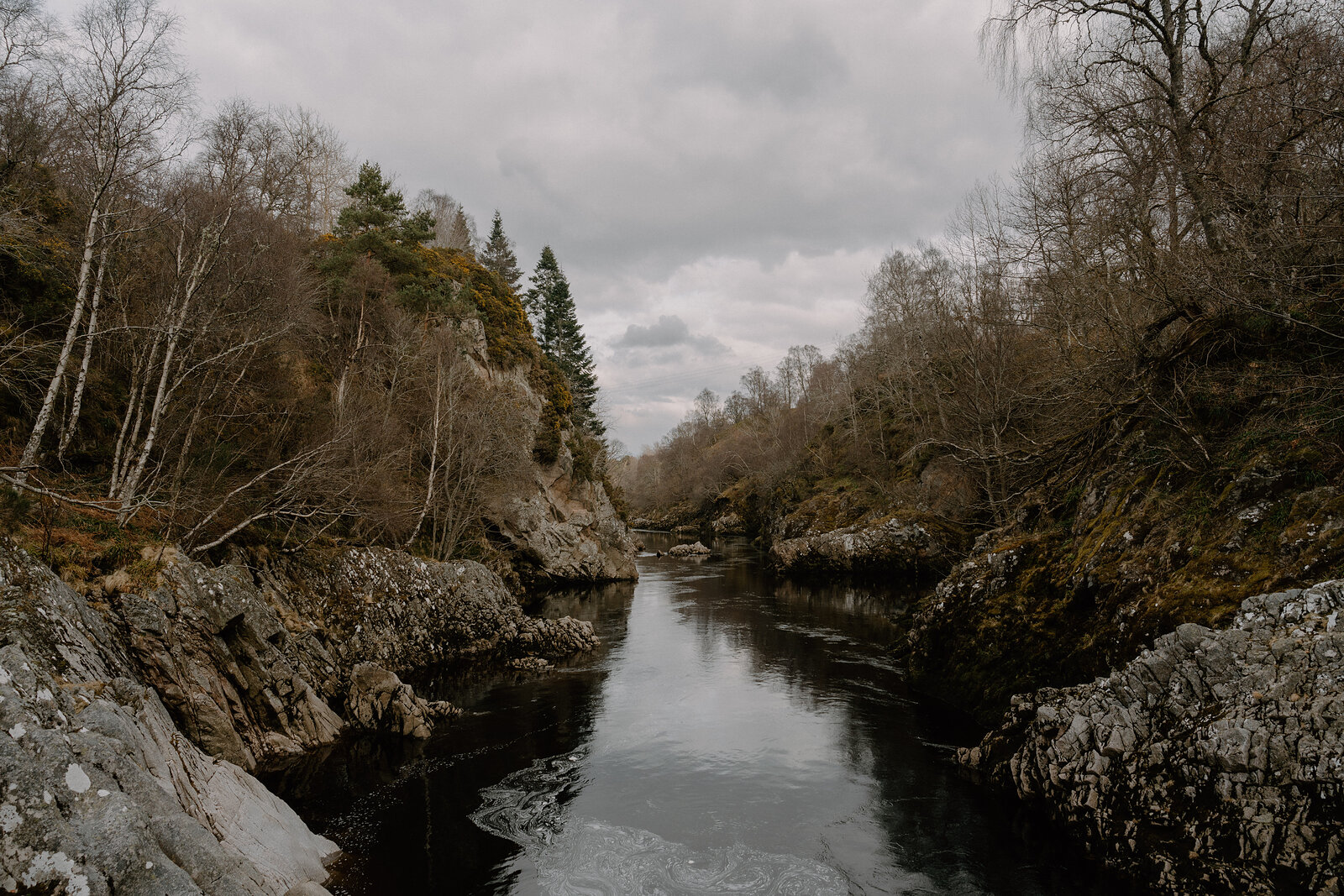 Bare trees line a rocky river gorge in Moray, the water running dark and still.