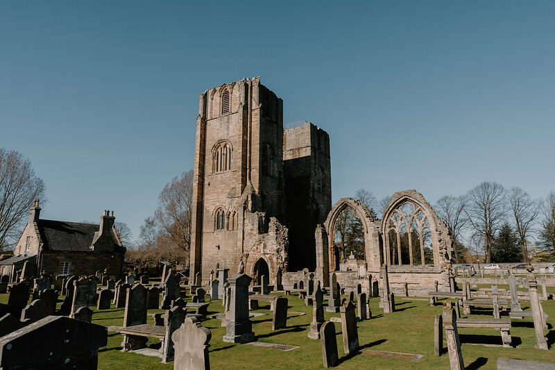 Elgin Cathedral's arched ruins and bell tower overlook a graveyard on a bright day.