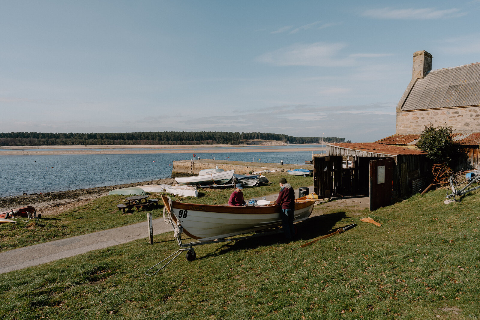 Two men work on a wooden rowing boat numbered 98 on the grass beside a boathouse on a calm Moray bay.