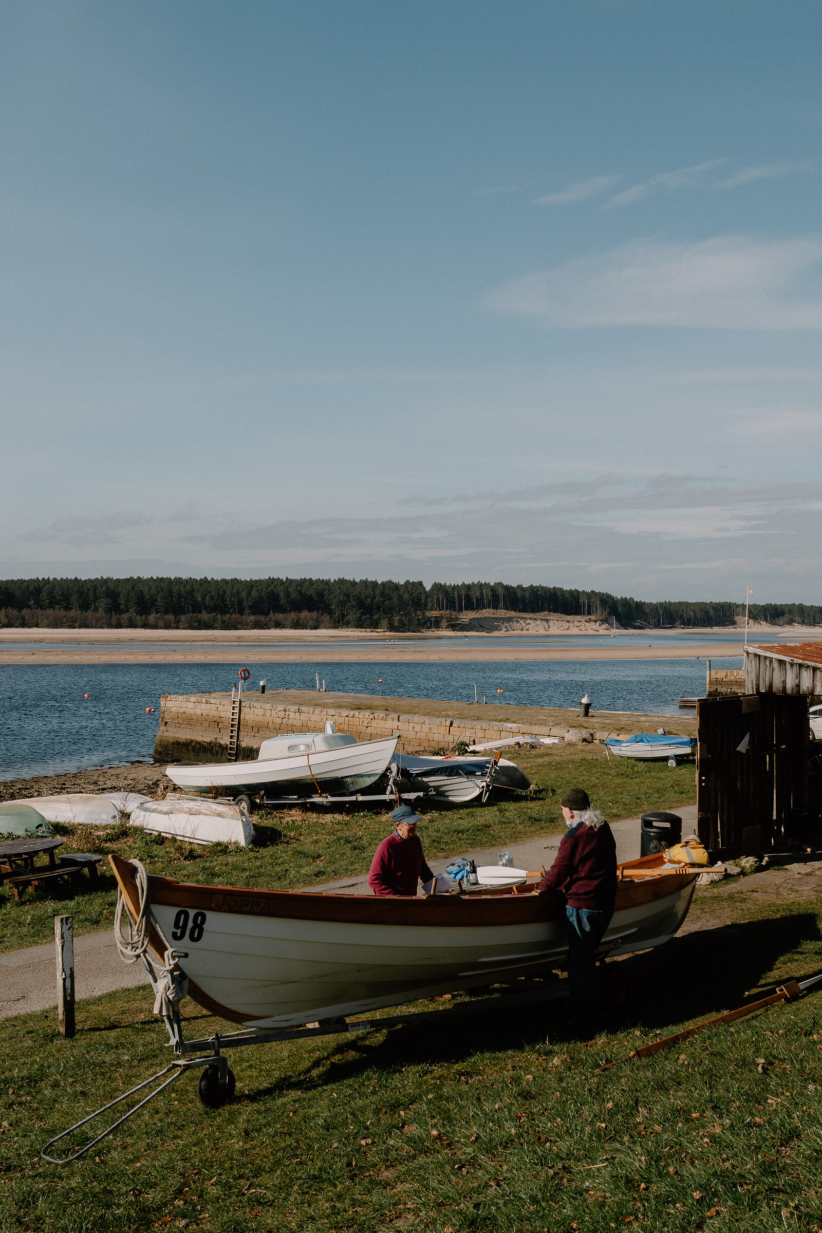 A wooden rowing boat numbered 98 sits on the grass at a boathouse beside a calm bay in Moray, two men working nearby.