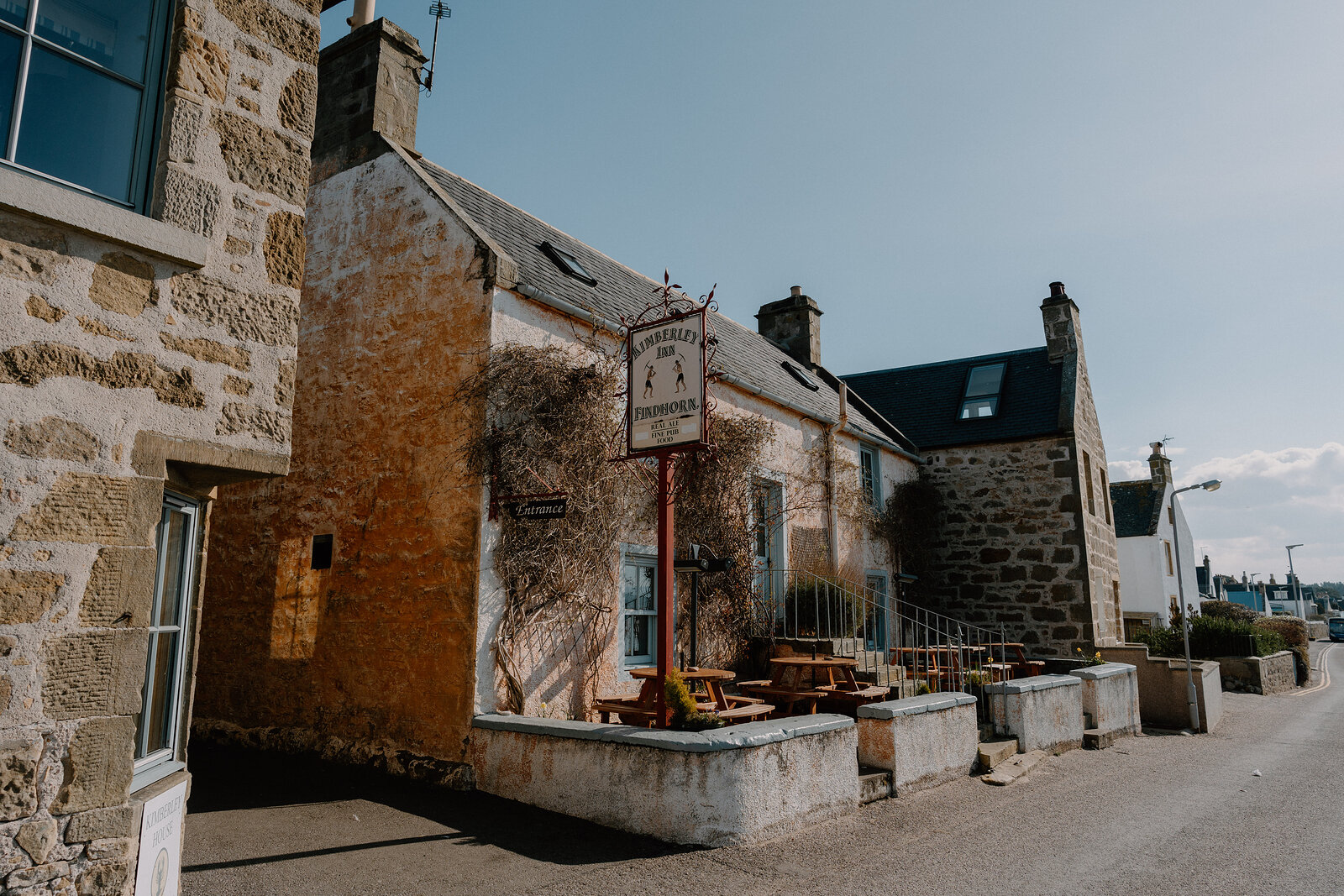 A stone cottage with a hanging pub sign and picnic tables sits on a Moray street corner, ivy climbing its walls.