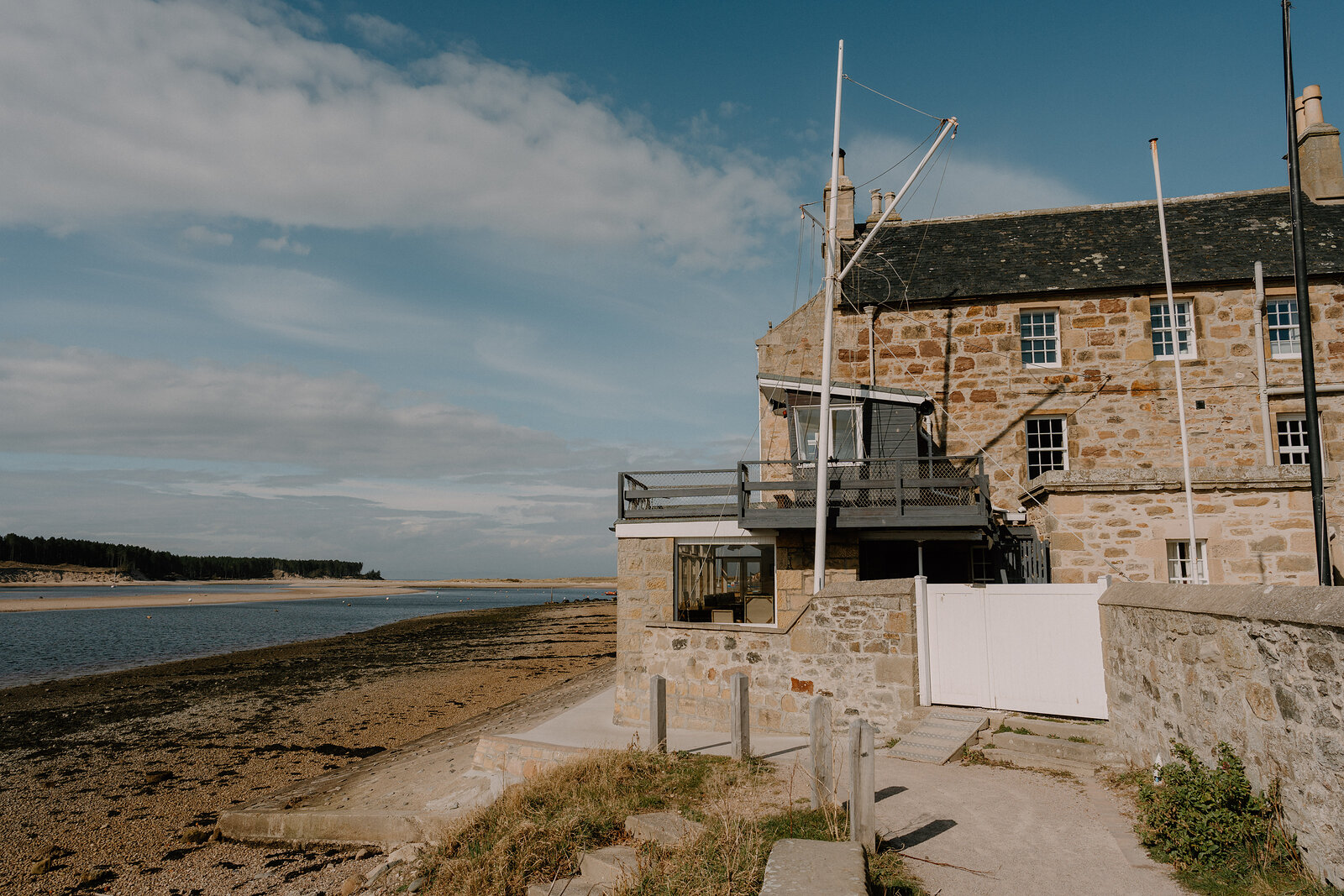 A stone building with a flagpole and mast stands above a sandy Moray bay.