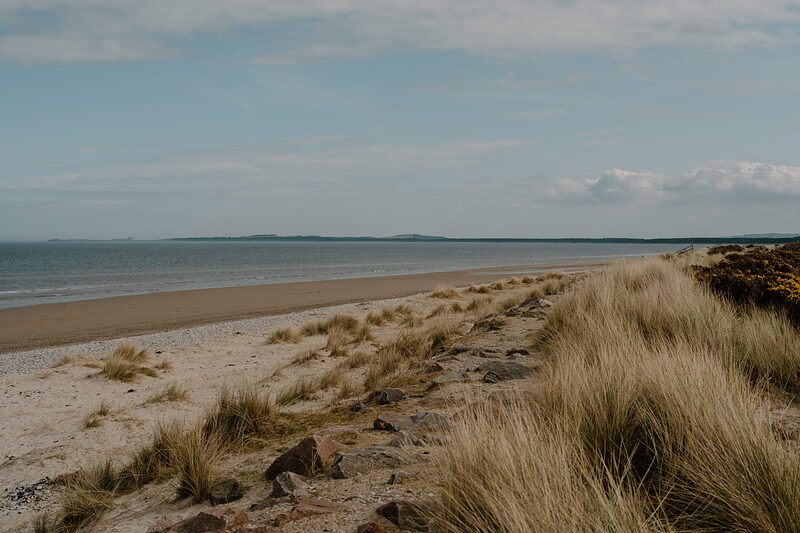 Dune grass frames a wide sandy Moray beach curving toward a distant headland.
