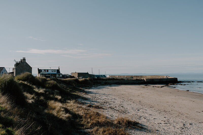 A row of harbourside cottages overlooks a sandy beach and stone pier in a Moray coastal village.