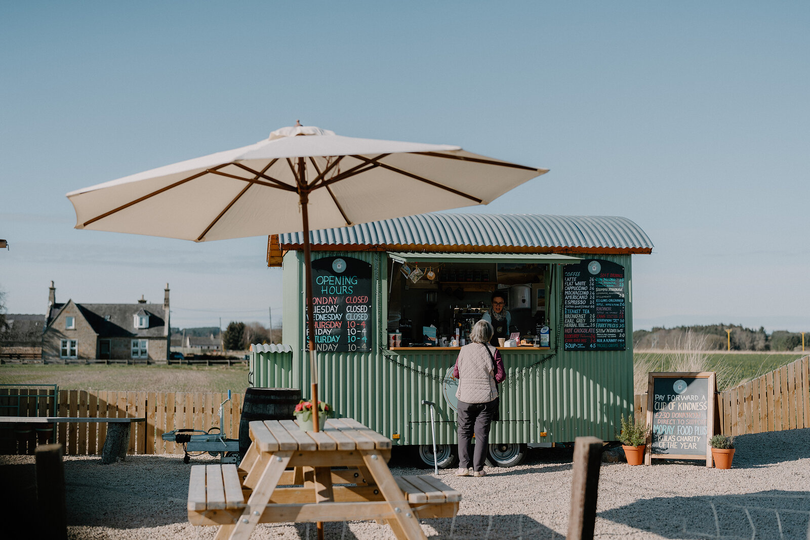 A customer orders at a green shepherd-hut coffee trailer beneath a large parasol in Moray.