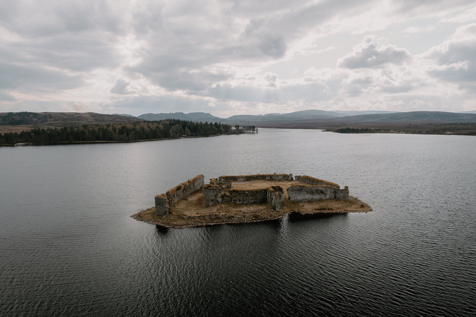 Lochindorb Castle's ruined walls sit surrounded by water, forested hills beyond, Moray.