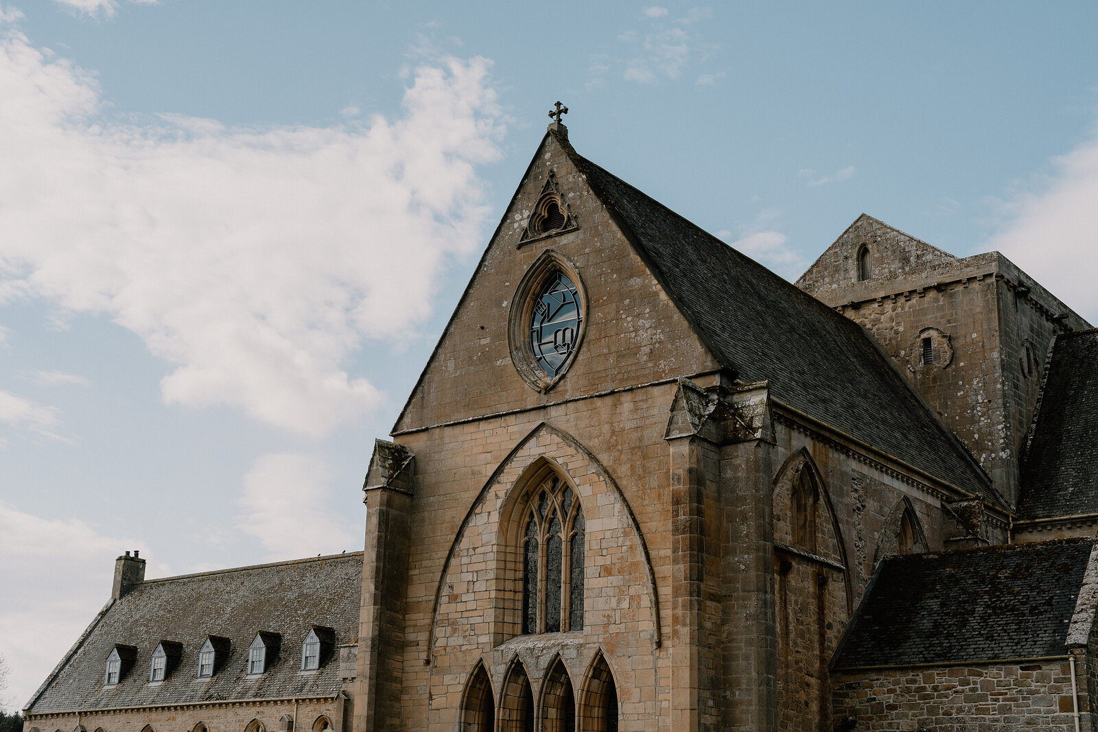 Pluscarden Abbey's arched windows and rose window rise beneath a stone cross against a blue sky.