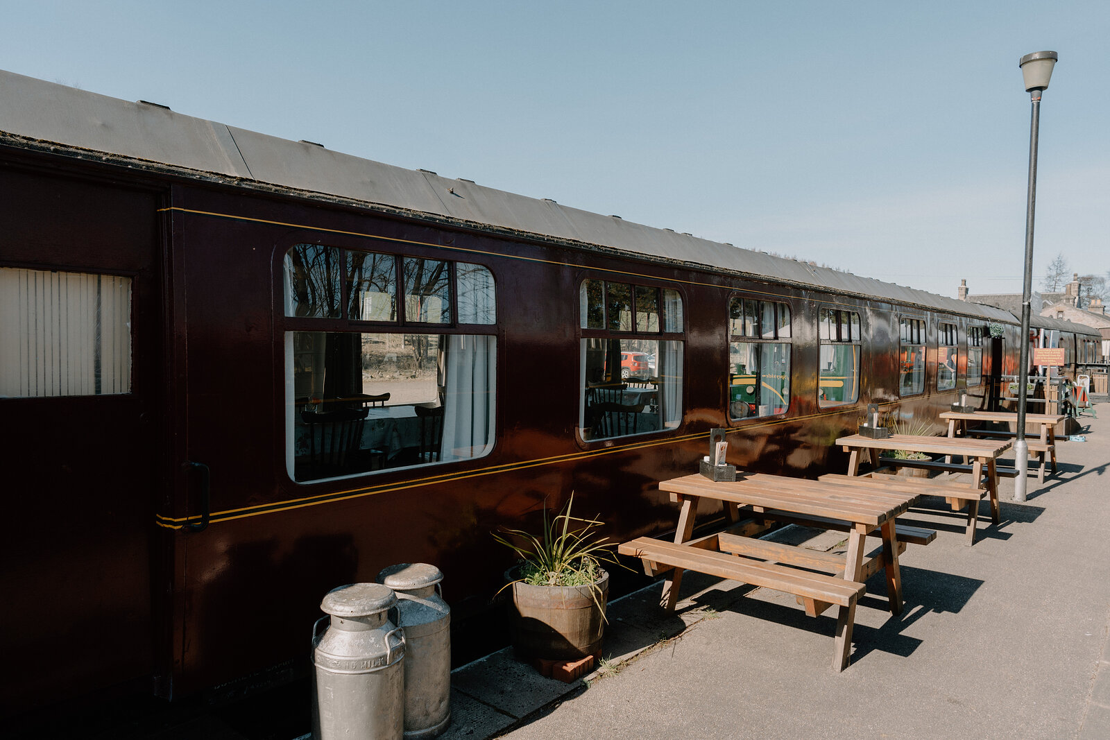 Sidings Café's maroon carriage sits behind milk churns and a potted plant on a sunny platform.