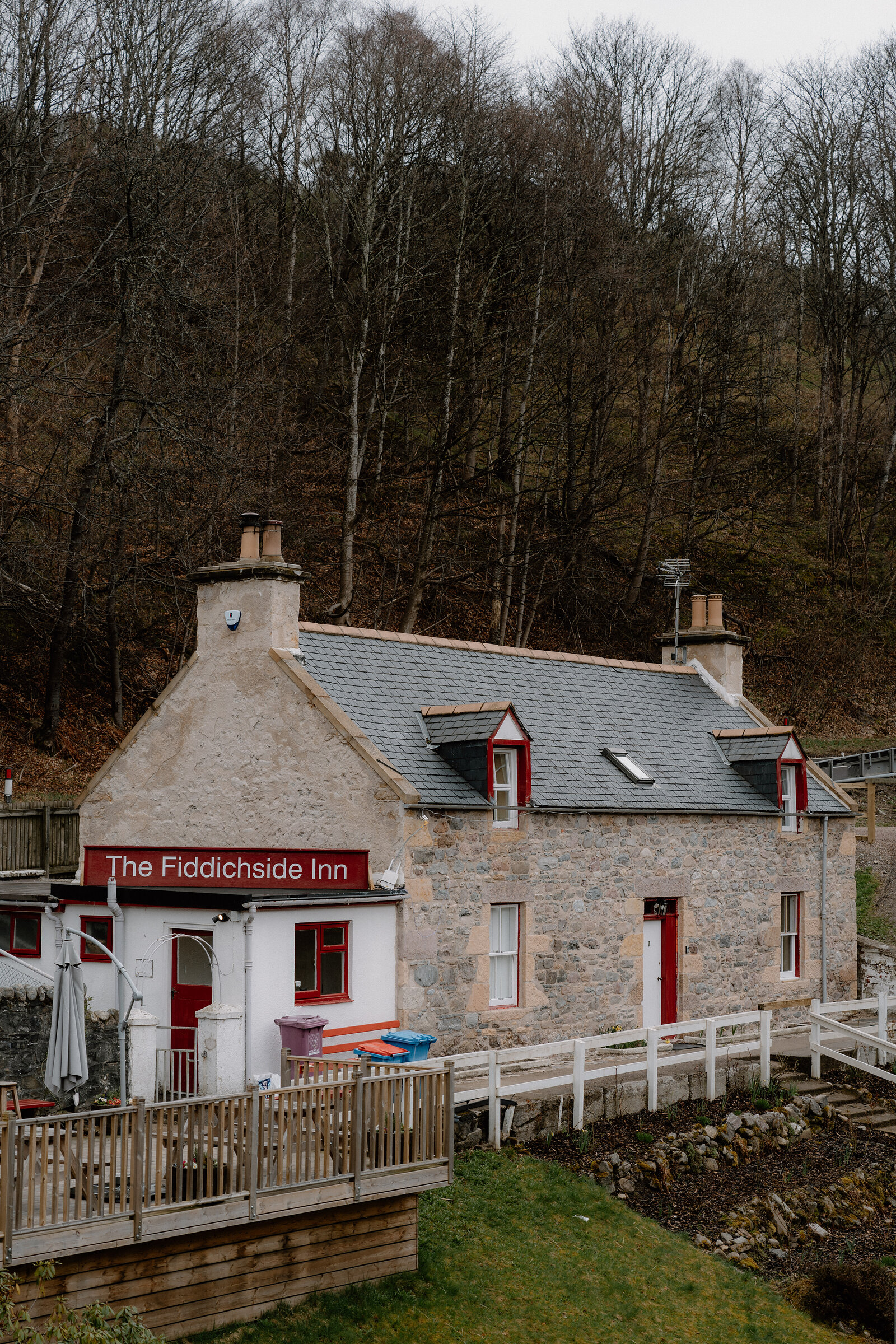 The Fiddichside Inn's stone and white-harled building sits among bare trees beside a wooden deck.