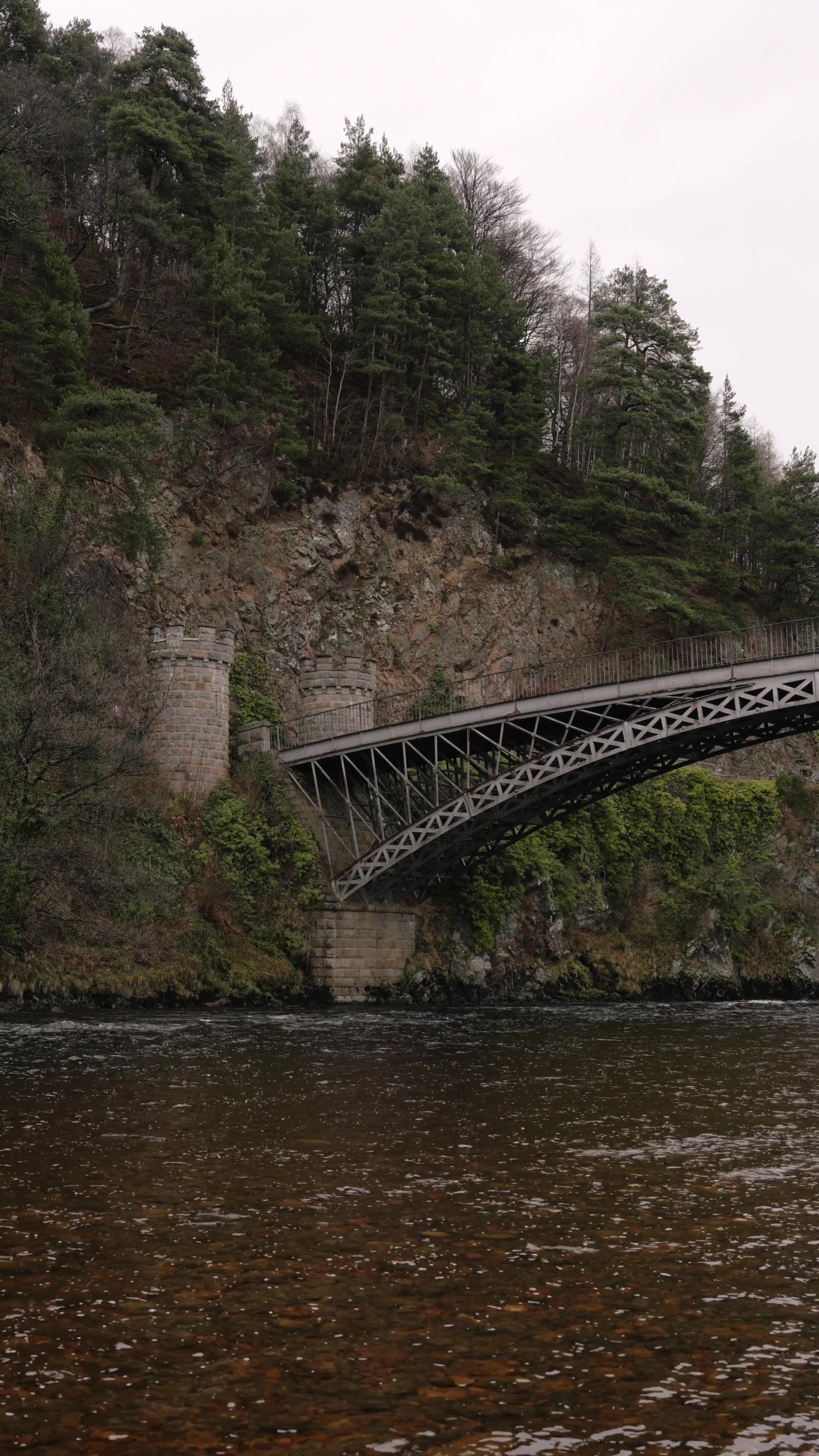 A cast-iron arched bridge rises between twin castellated stone towers over a rocky river, pine-covered slopes framing the crossing.