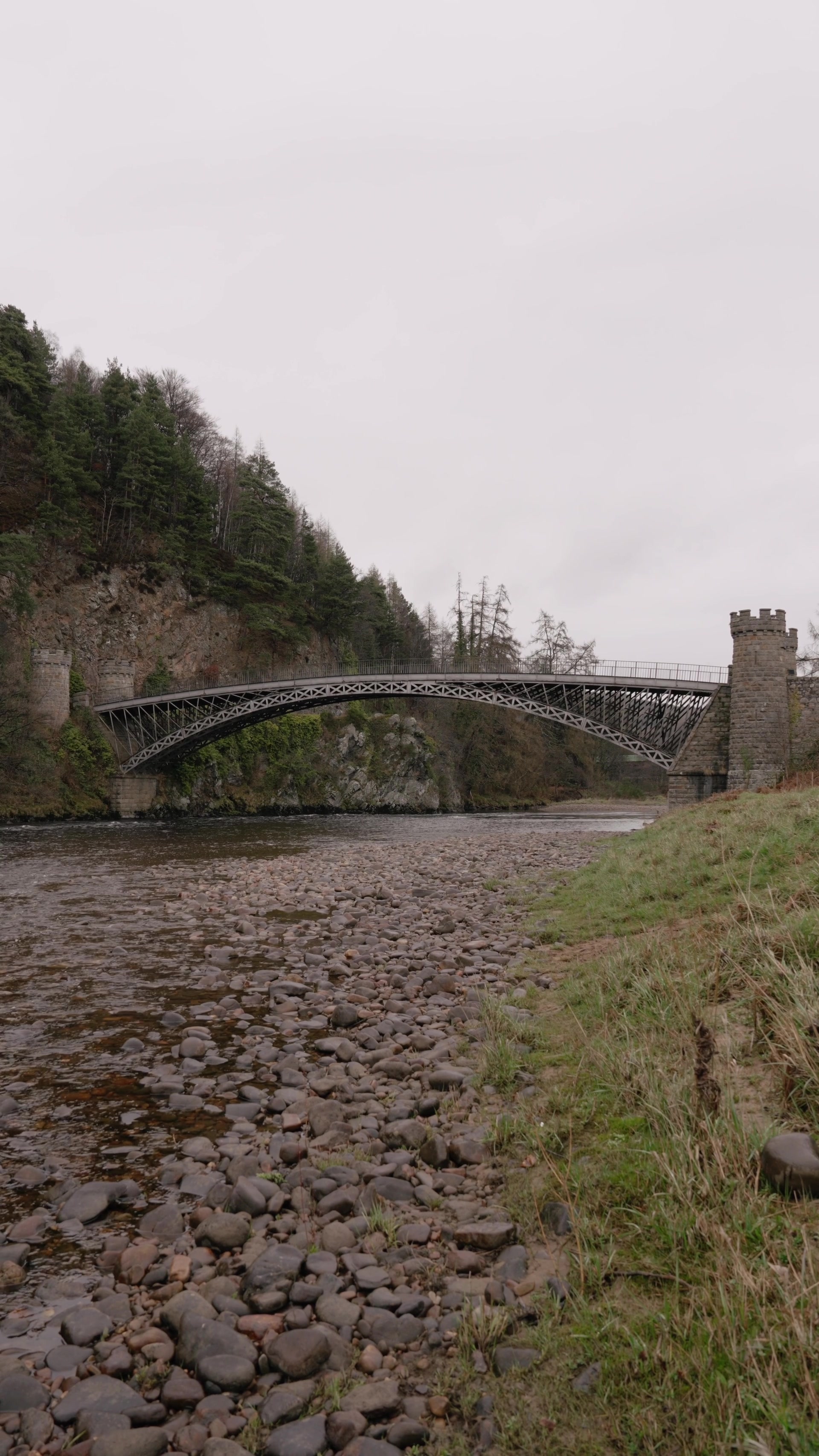 A cast-iron arched bridge with castellated stone towers spans the River Spey beside a wooded cliff, a pebble riverbank stretching along the near shore.