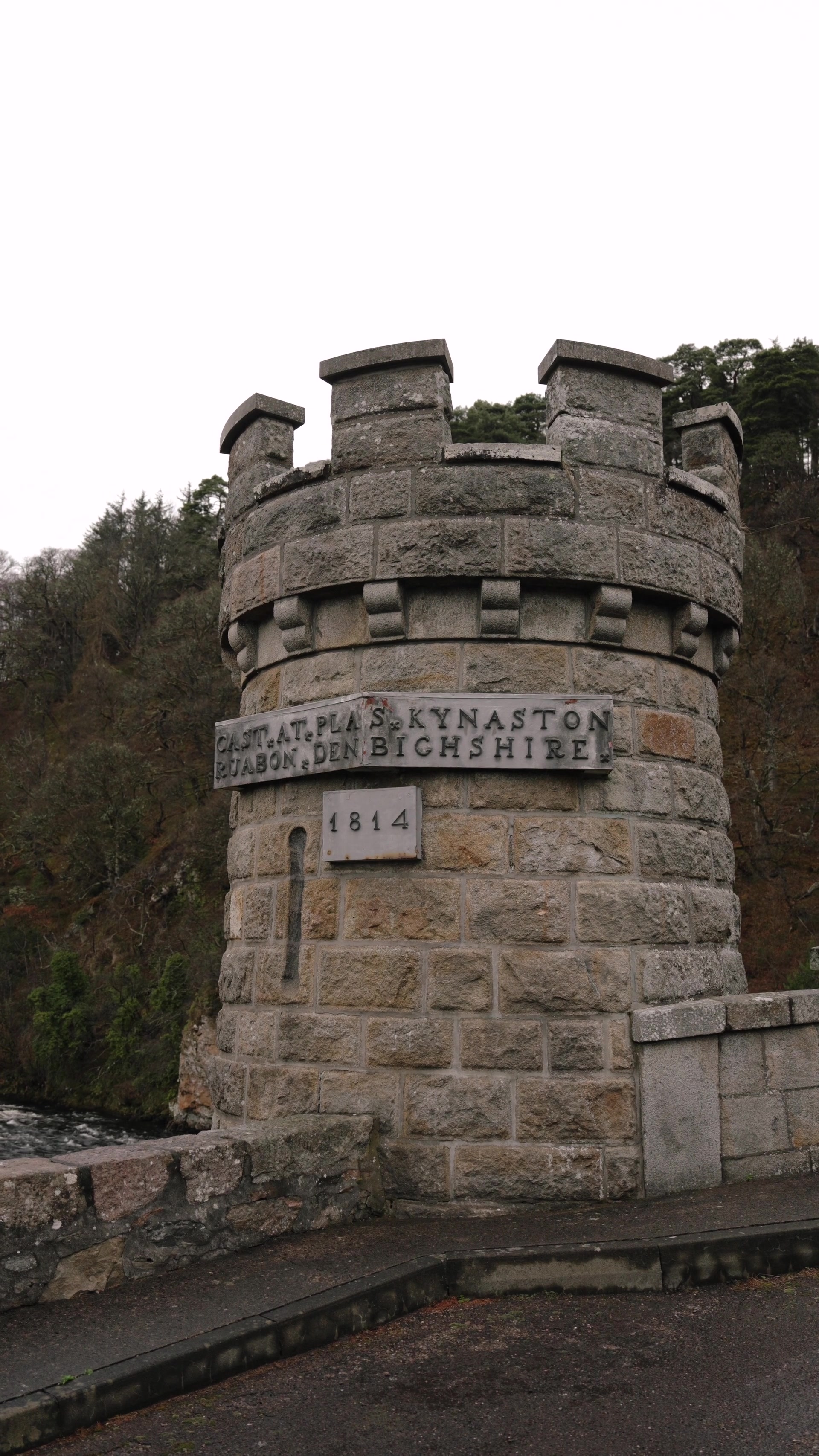 A weathered stone tower with battlements bears a plaque reading "Cast at Plas Kynaston, Ruabon, Denbighshire" and a date stone marked "1814", pine trees rising on the wooded bank behind.