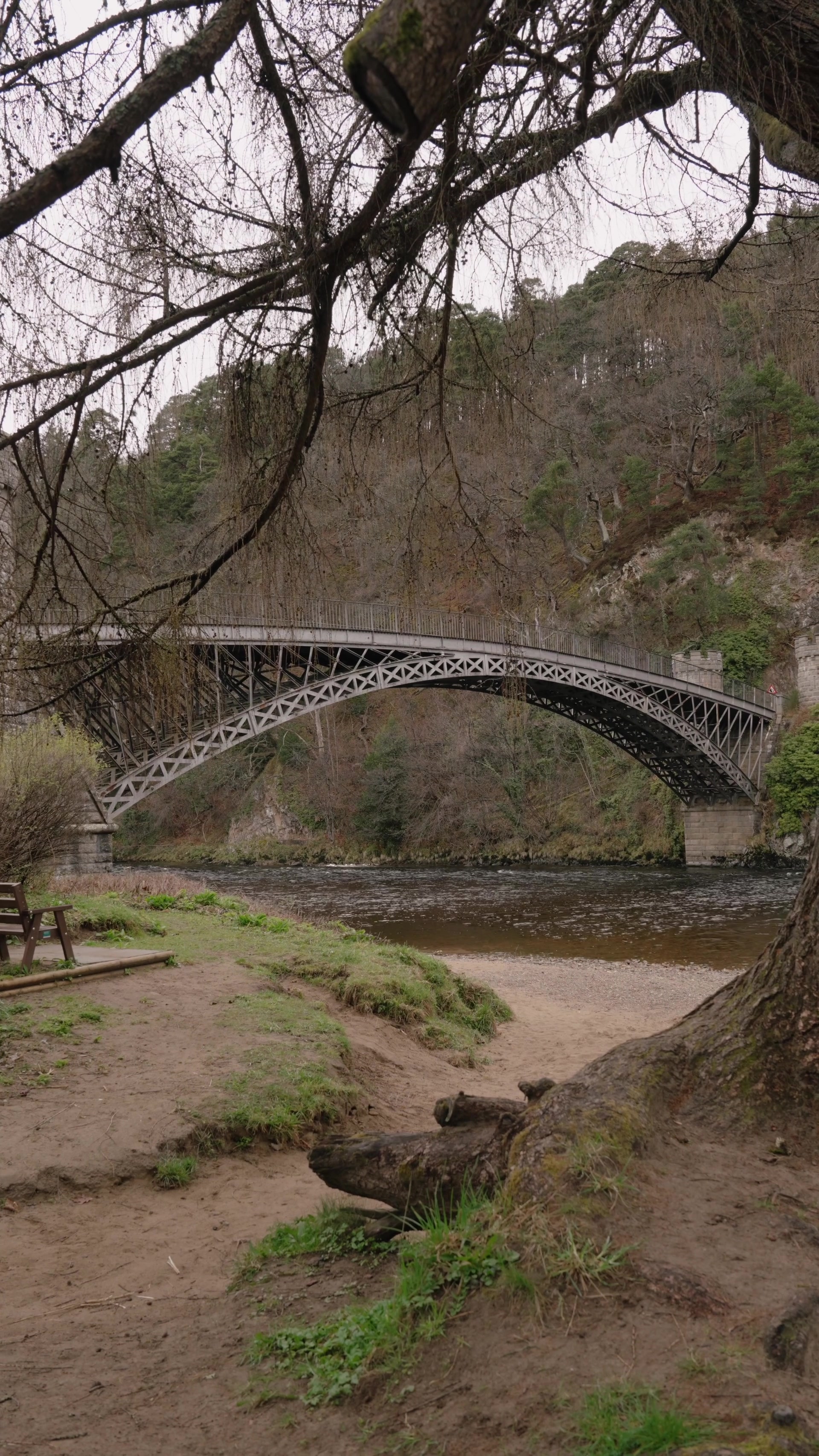 A cast-iron arched bridge spans a wide river between wooded cliffs, a sandy riverbank and picnic bench visible beneath overhanging branches in the foreground.